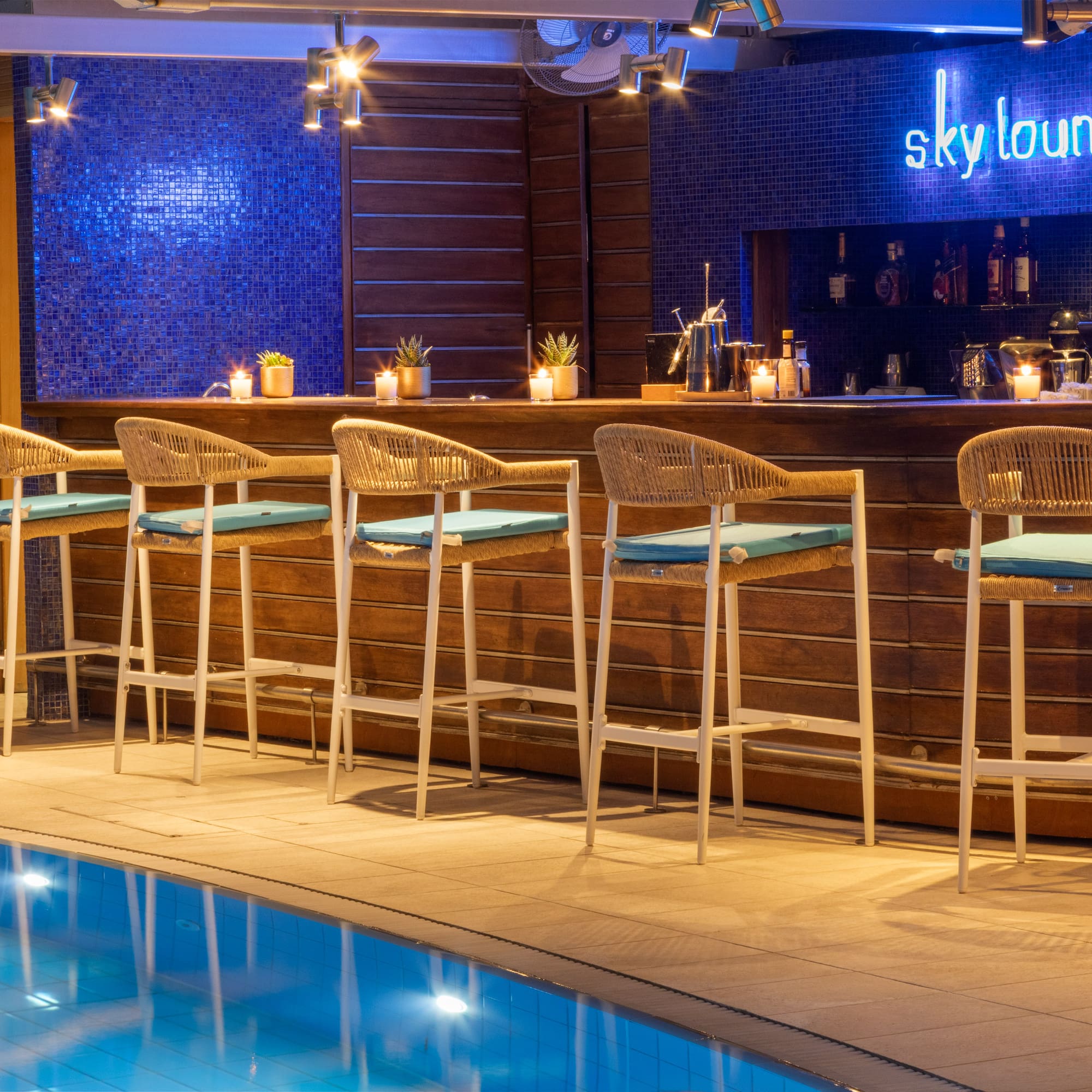Hotel Sky Lounge bar with neon sign, pool, and wicker stools at night.