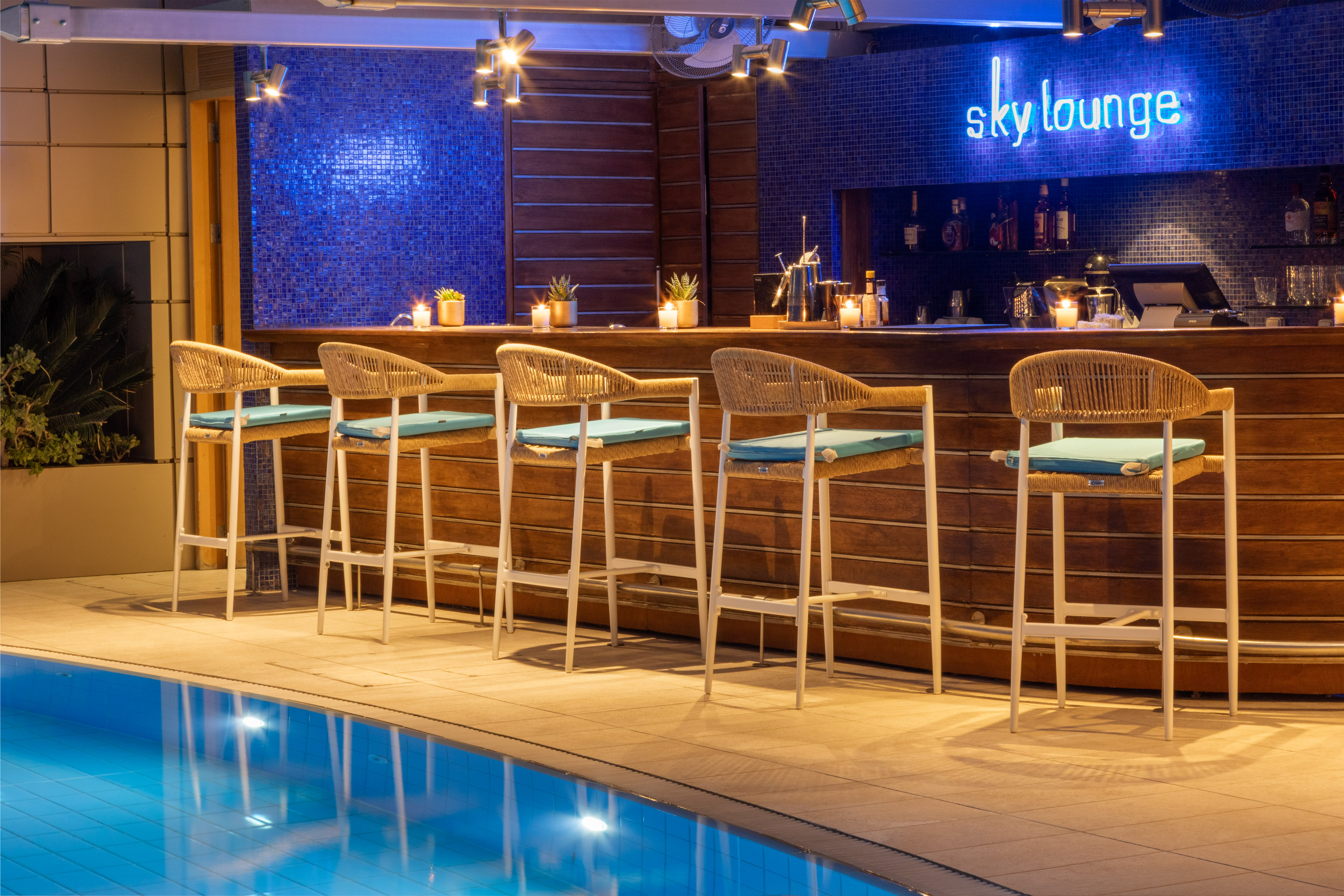 Hotel Sky Lounge bar with neon sign, pool, and wicker stools at night.