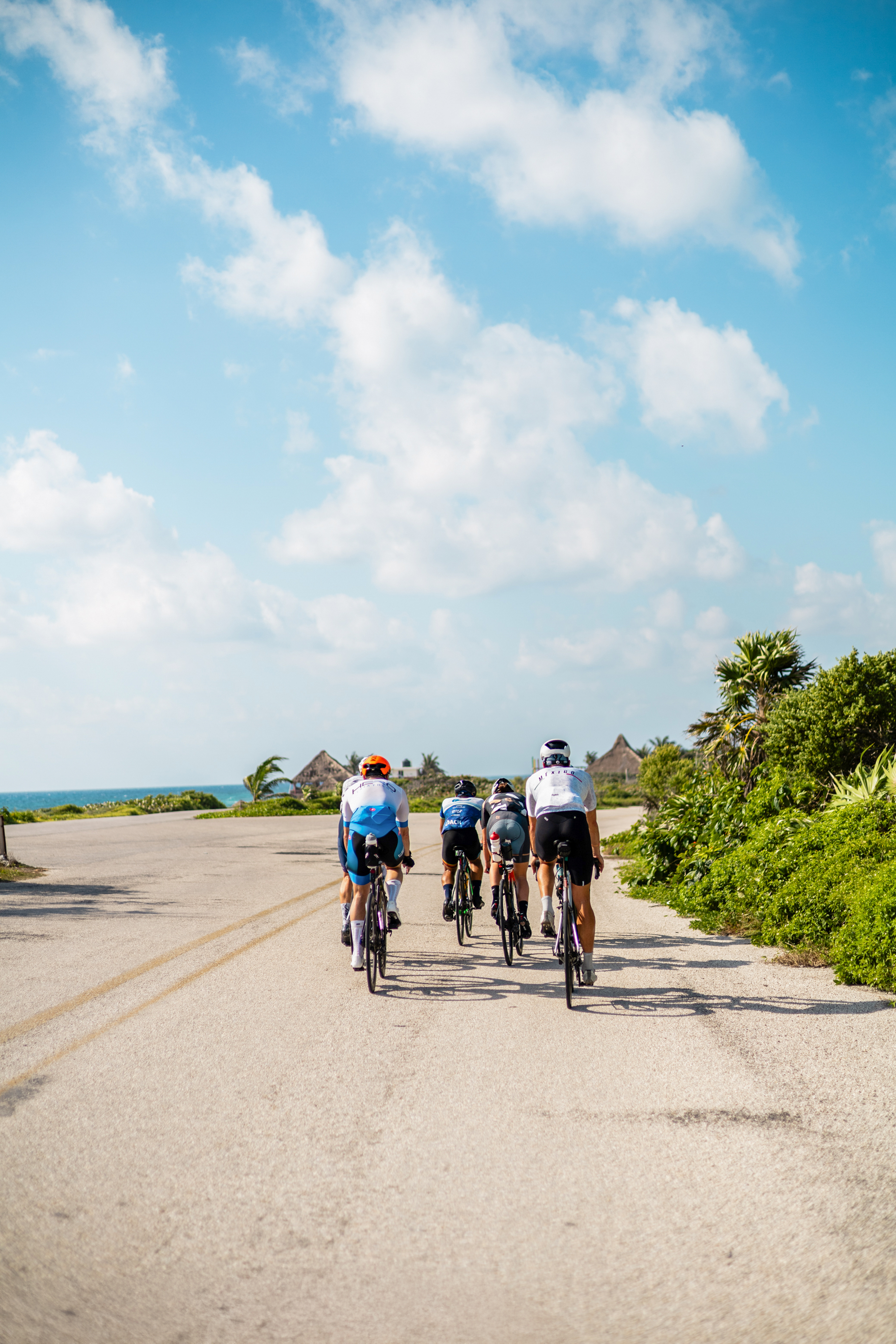 Cyclists on coastal road with ocean, tropical huts, and lush greenery.
