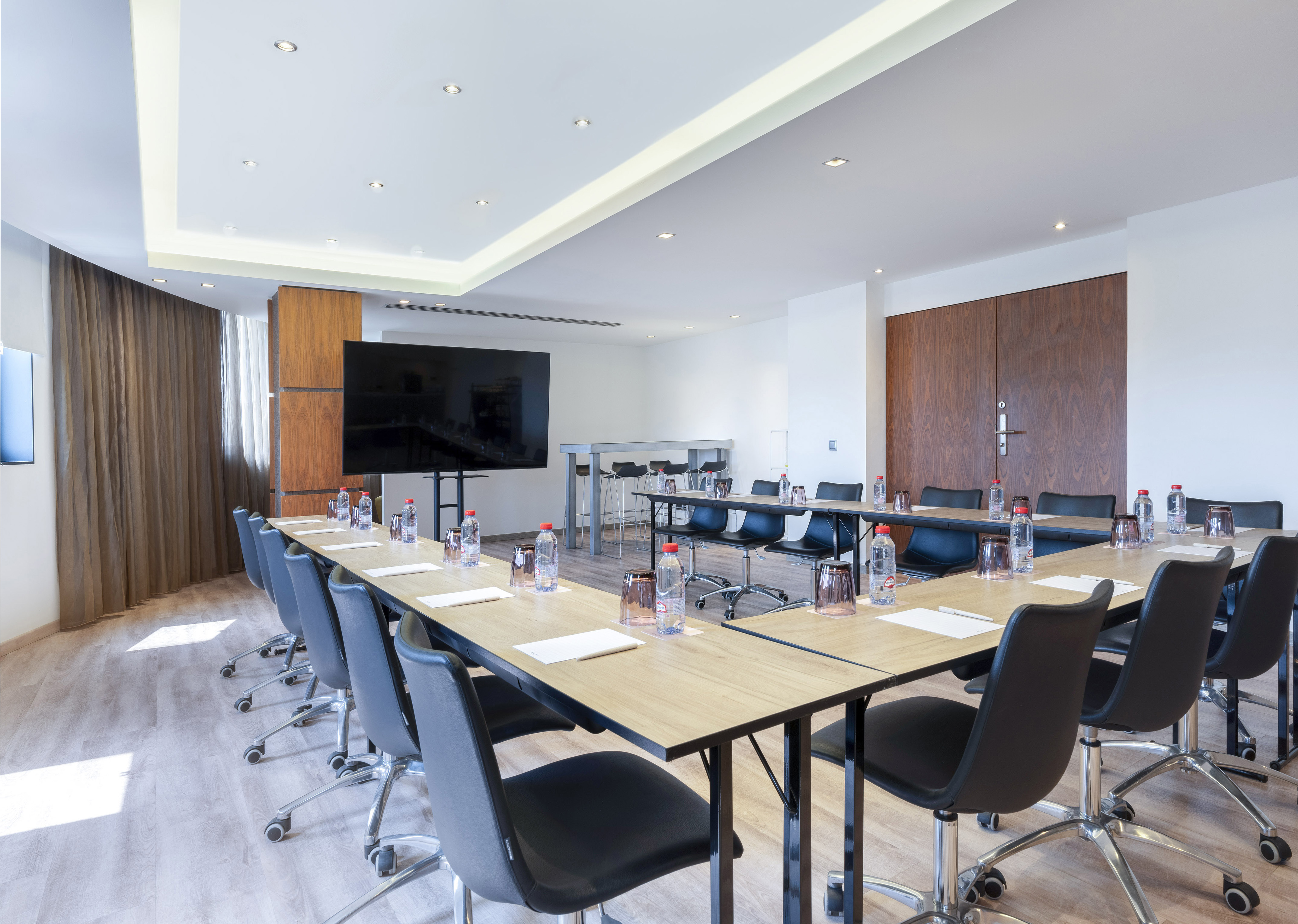 Modern U-shaped conference room with large TV, ergonomic chairs, water bottles.