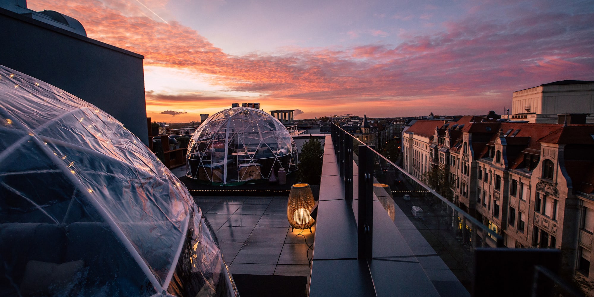 Rooftop bar with transparent dining igloos overlooking a city sunset.