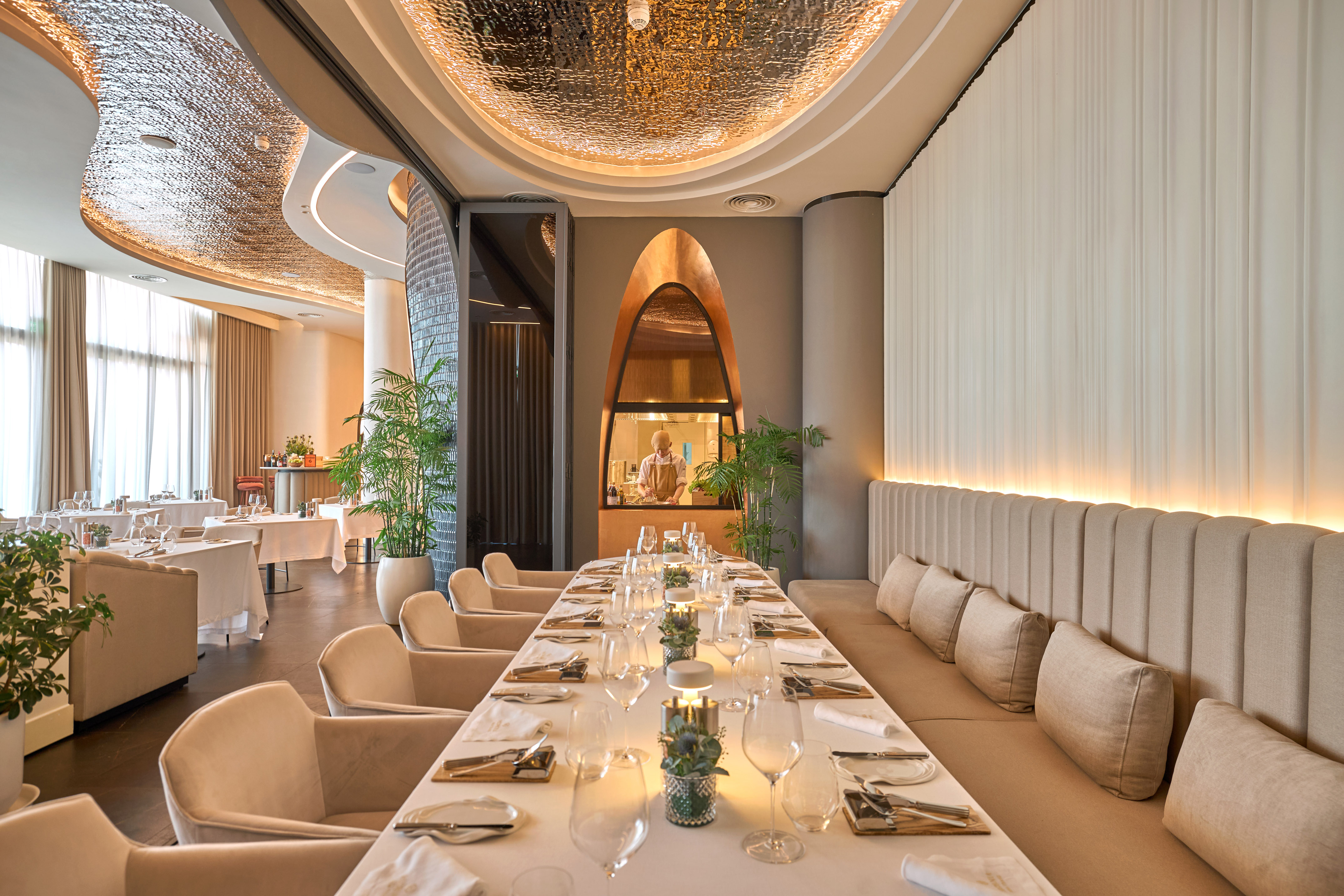 Elegant restaurant interior with long dining table, textured ceiling, and chef's kitchen.