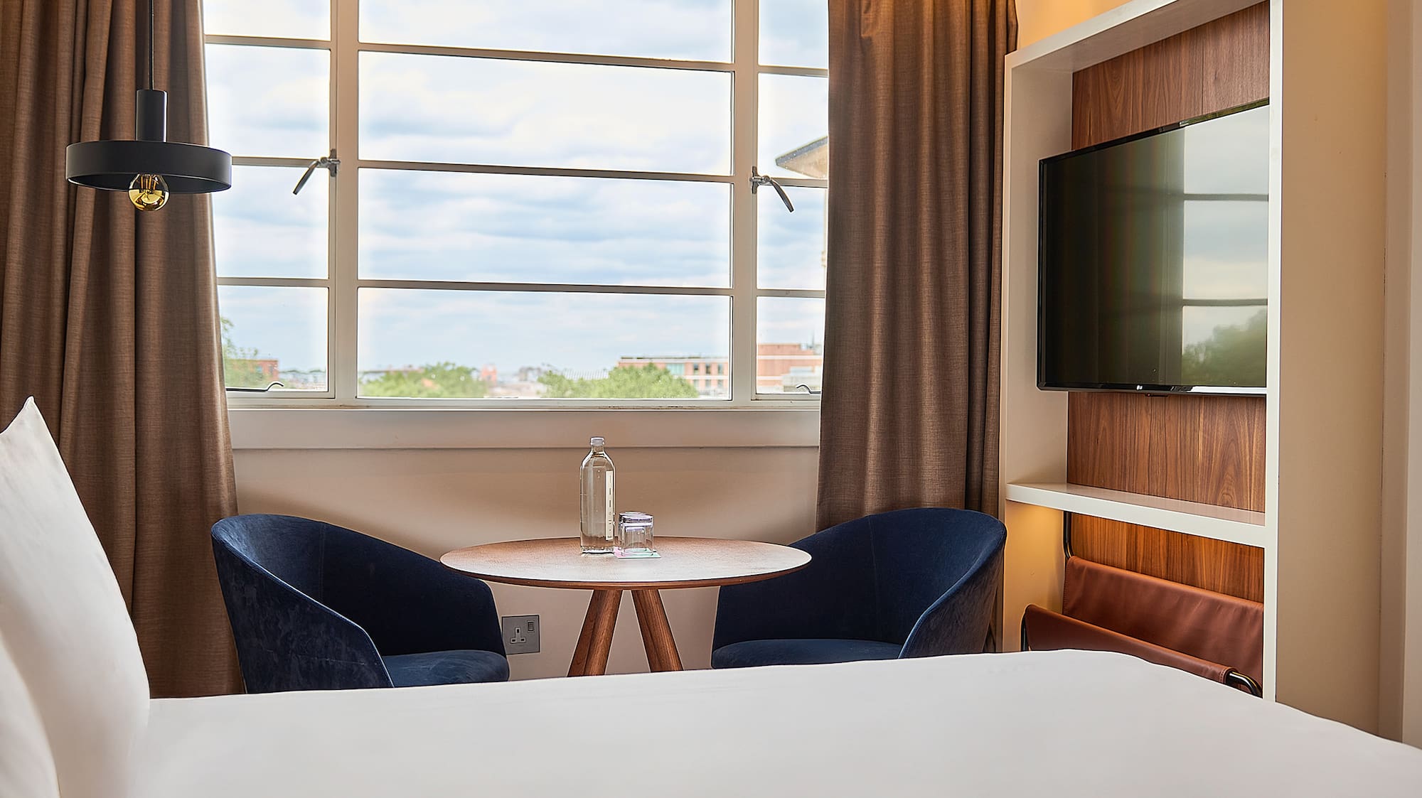 Contemporary hotel room: bed, window view, blue chairs, wooden table, and TV.