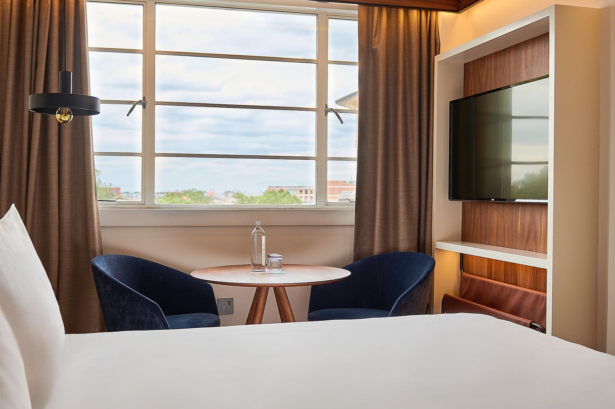 Contemporary hotel room: bed, window view, blue chairs, wooden table, and TV.