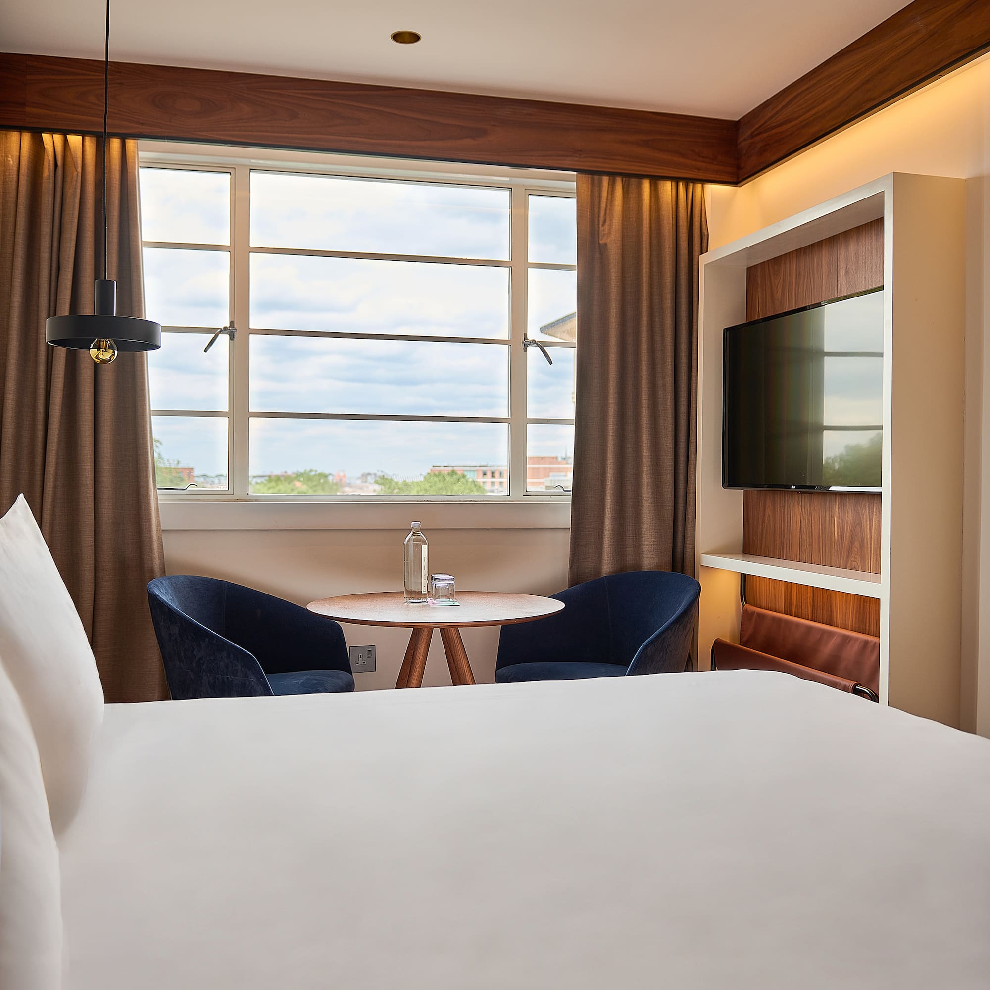Contemporary hotel room: bed, window view, blue chairs, wooden table, and TV.