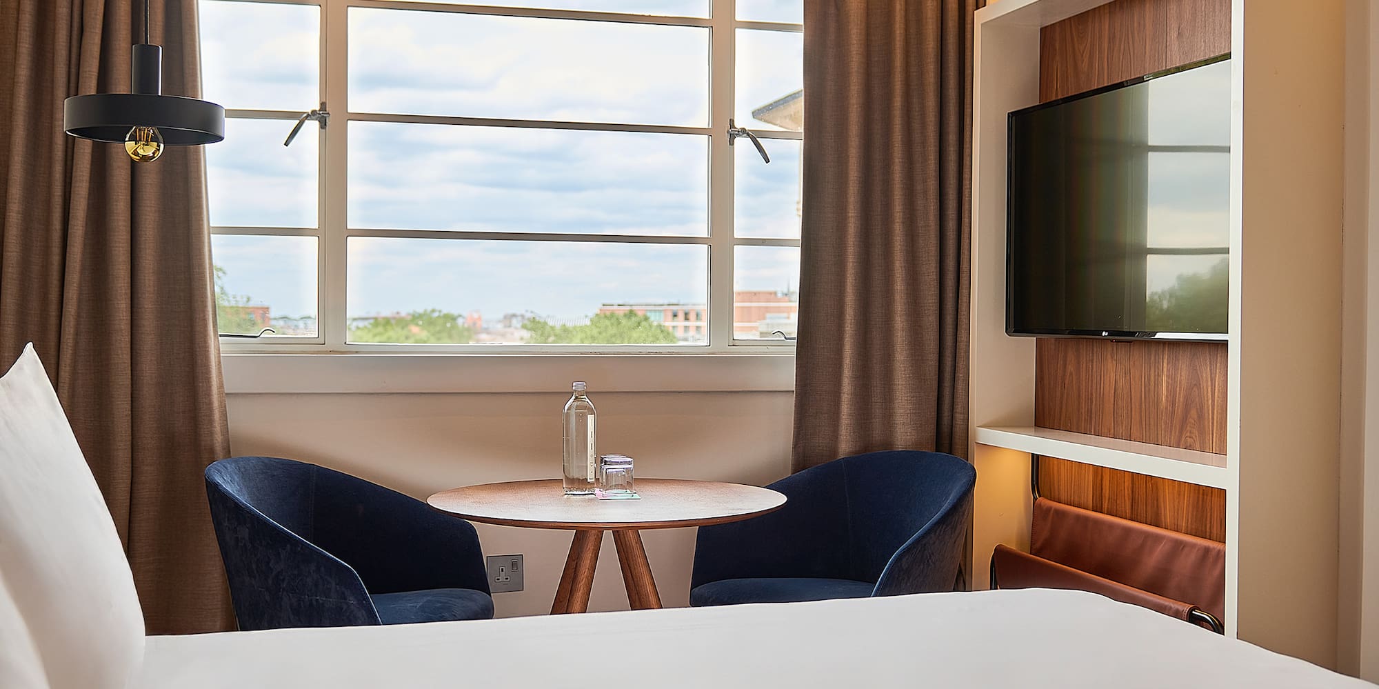 Contemporary hotel room: bed, window view, blue chairs, wooden table, and TV.