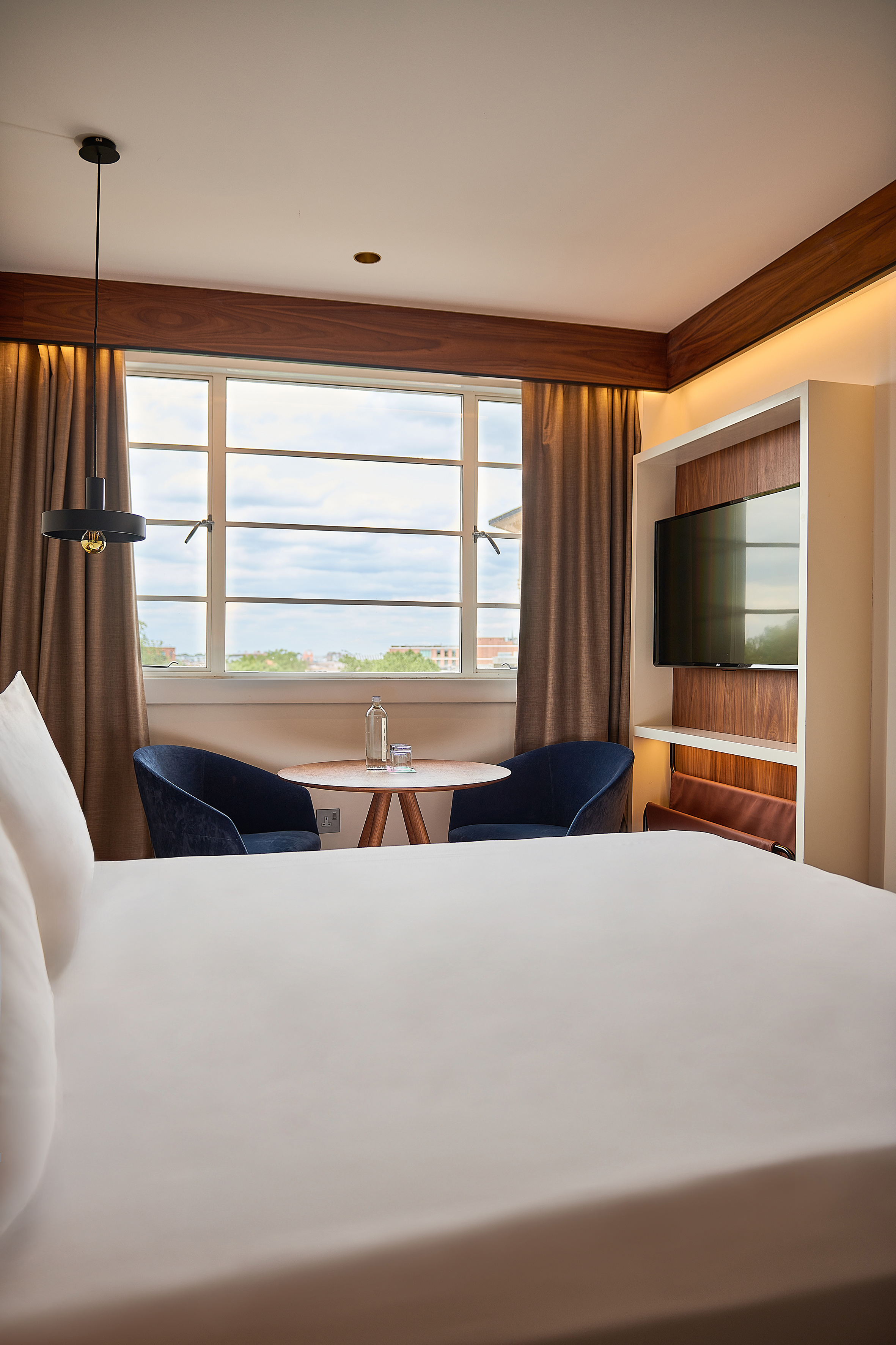 Contemporary hotel room: bed, window view, blue chairs, wooden table, and TV.
