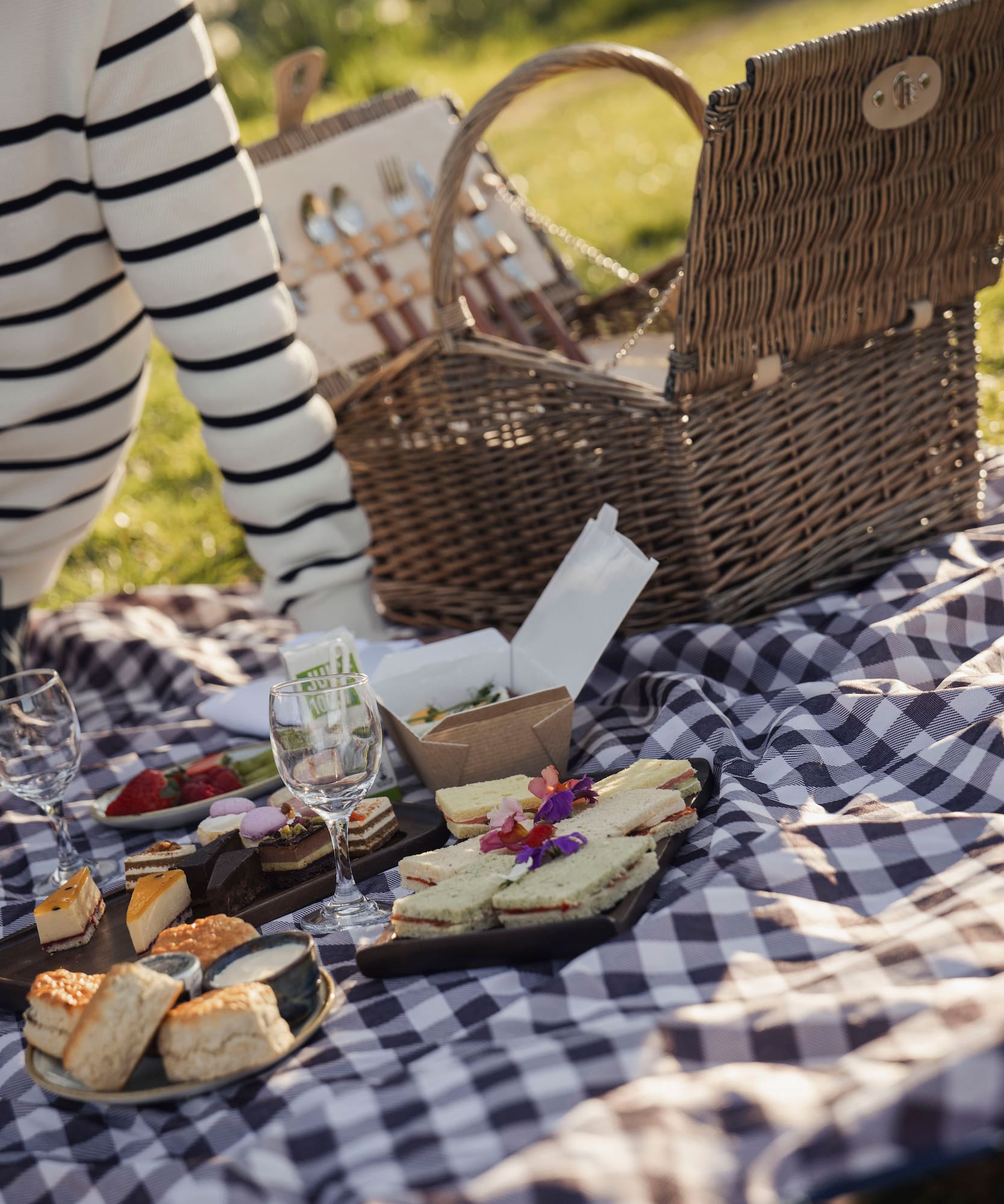 Picnic spread on checkered blanket: sandwiches, desserts, wine, wicker basket.