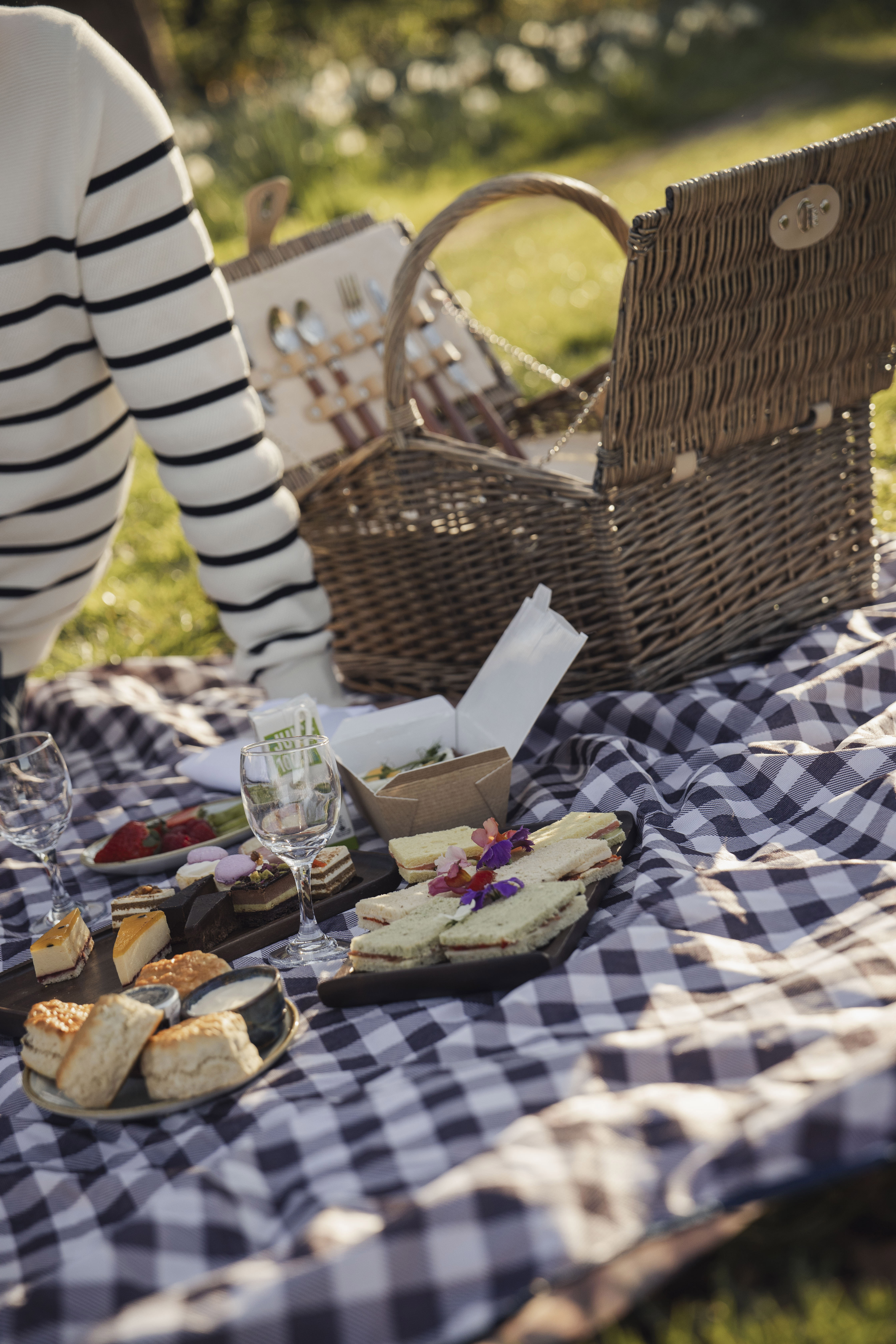 Picnic spread on checkered blanket: sandwiches, desserts, wine, wicker basket.