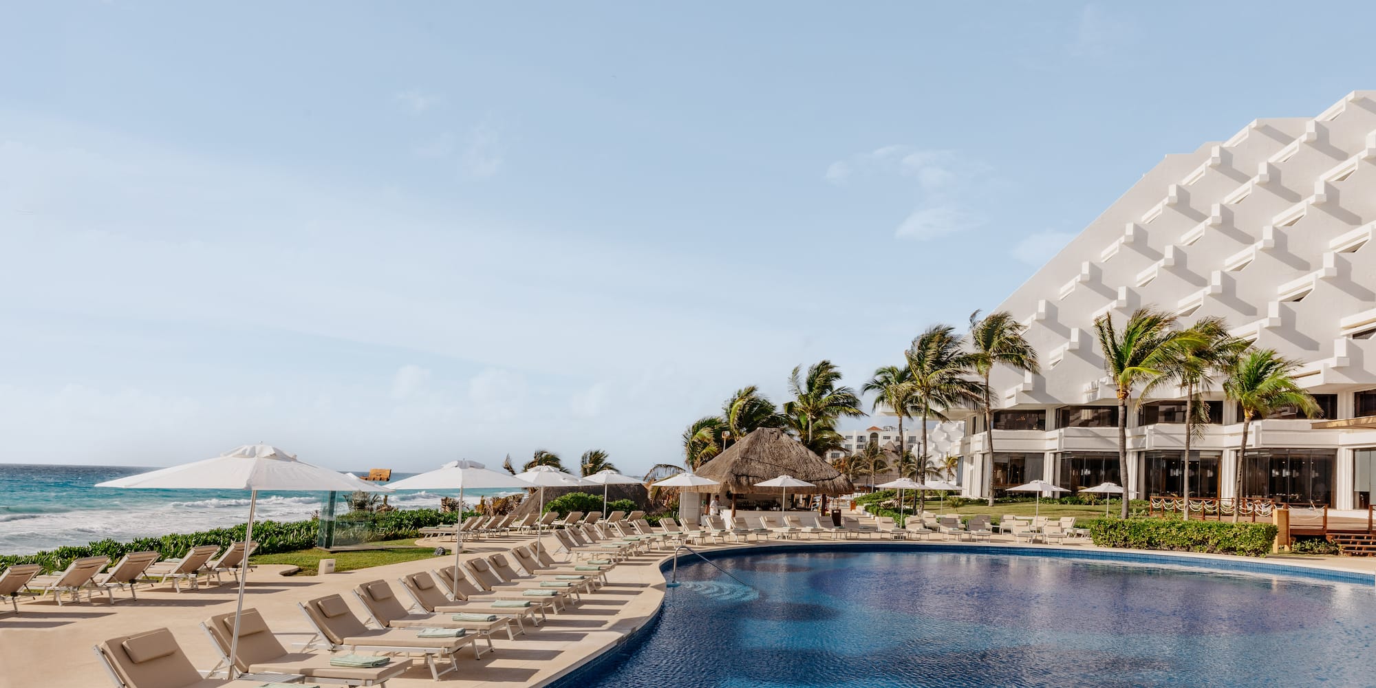Beach resort pool with lounge chairs, palm trees, and ocean view.