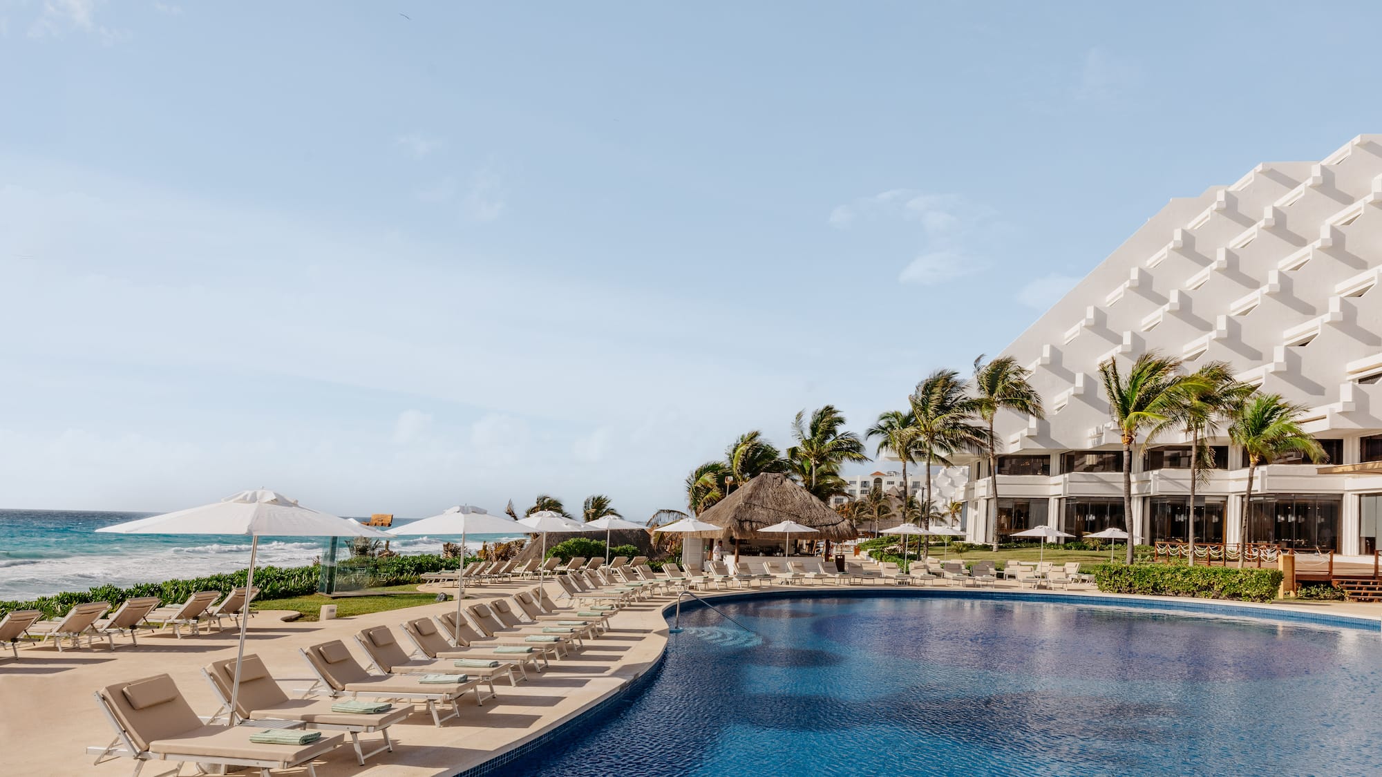 Beach resort pool with lounge chairs, palm trees, and ocean view.