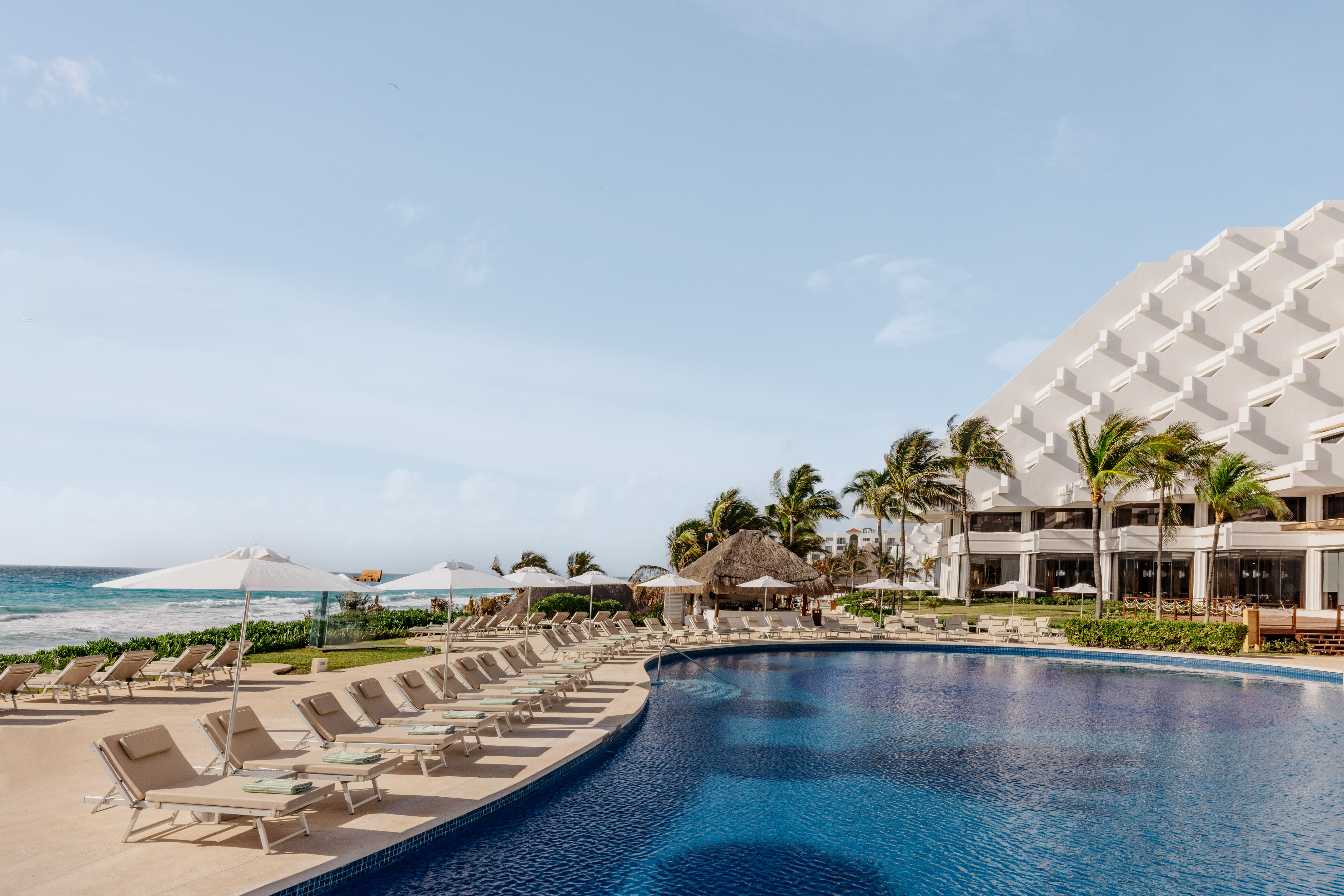 Beach resort pool with lounge chairs, palm trees, and ocean view.