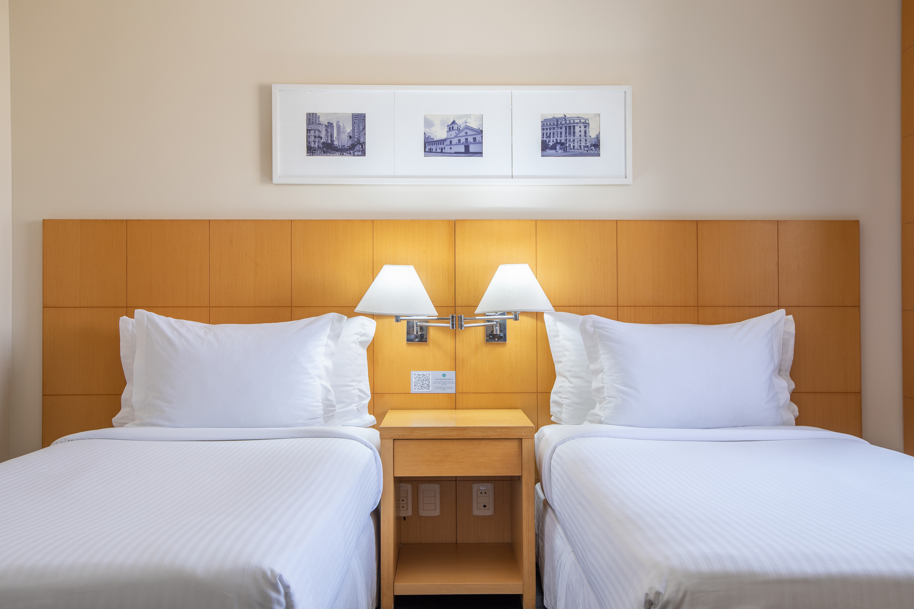 Hotel room with twin beds, wooden headboard, nightstand, lamps, and city artwork.