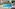 Curved resort pool with sunbeds, straw umbrellas, and hotel complex.