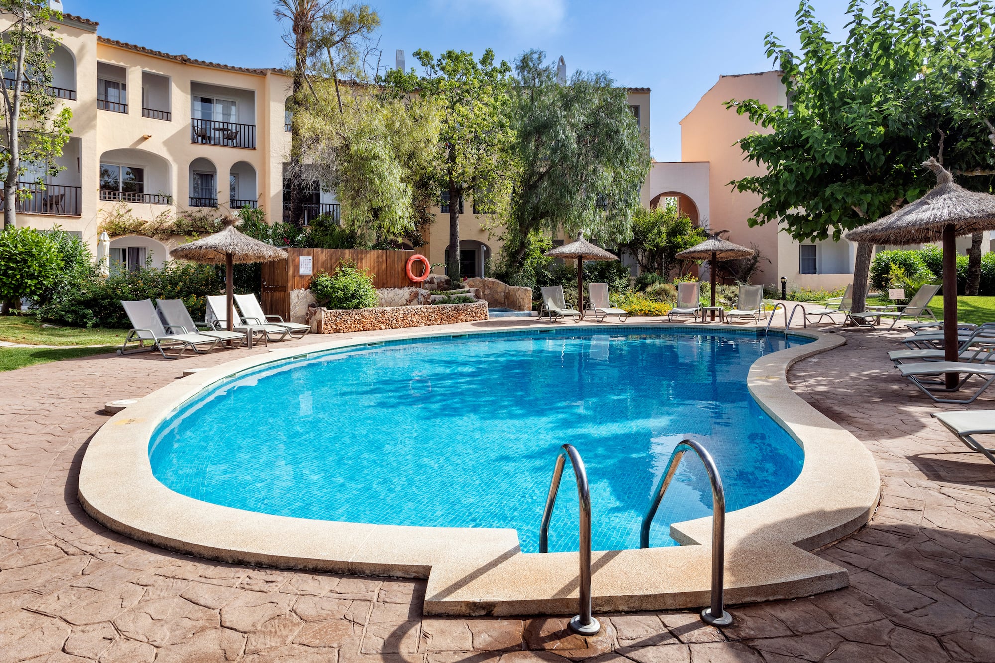 Curved resort pool with sunbeds, straw umbrellas, and hotel complex.