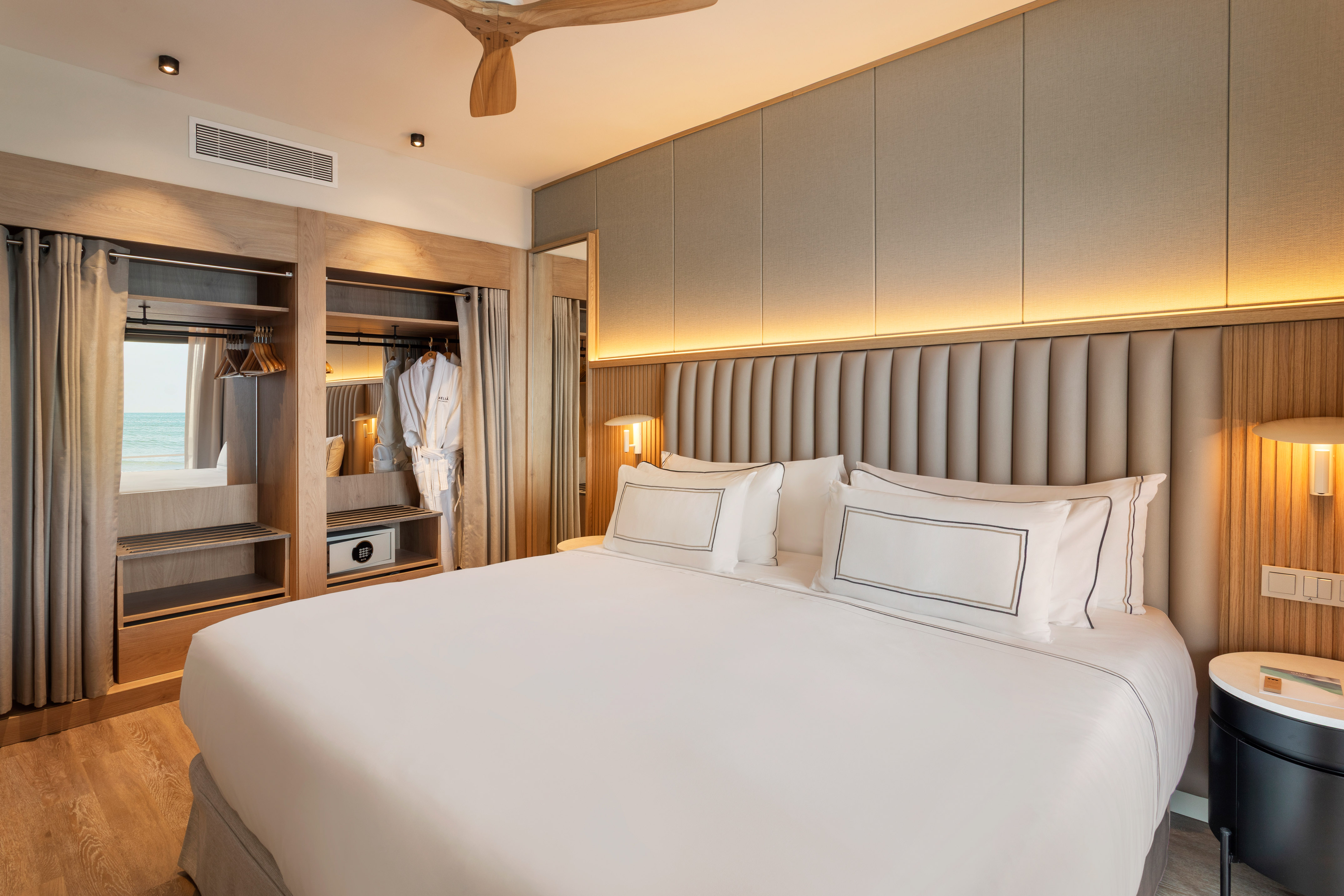 Modern hotel bedroom with king bed, ocean view, open wardrobe, and safe.