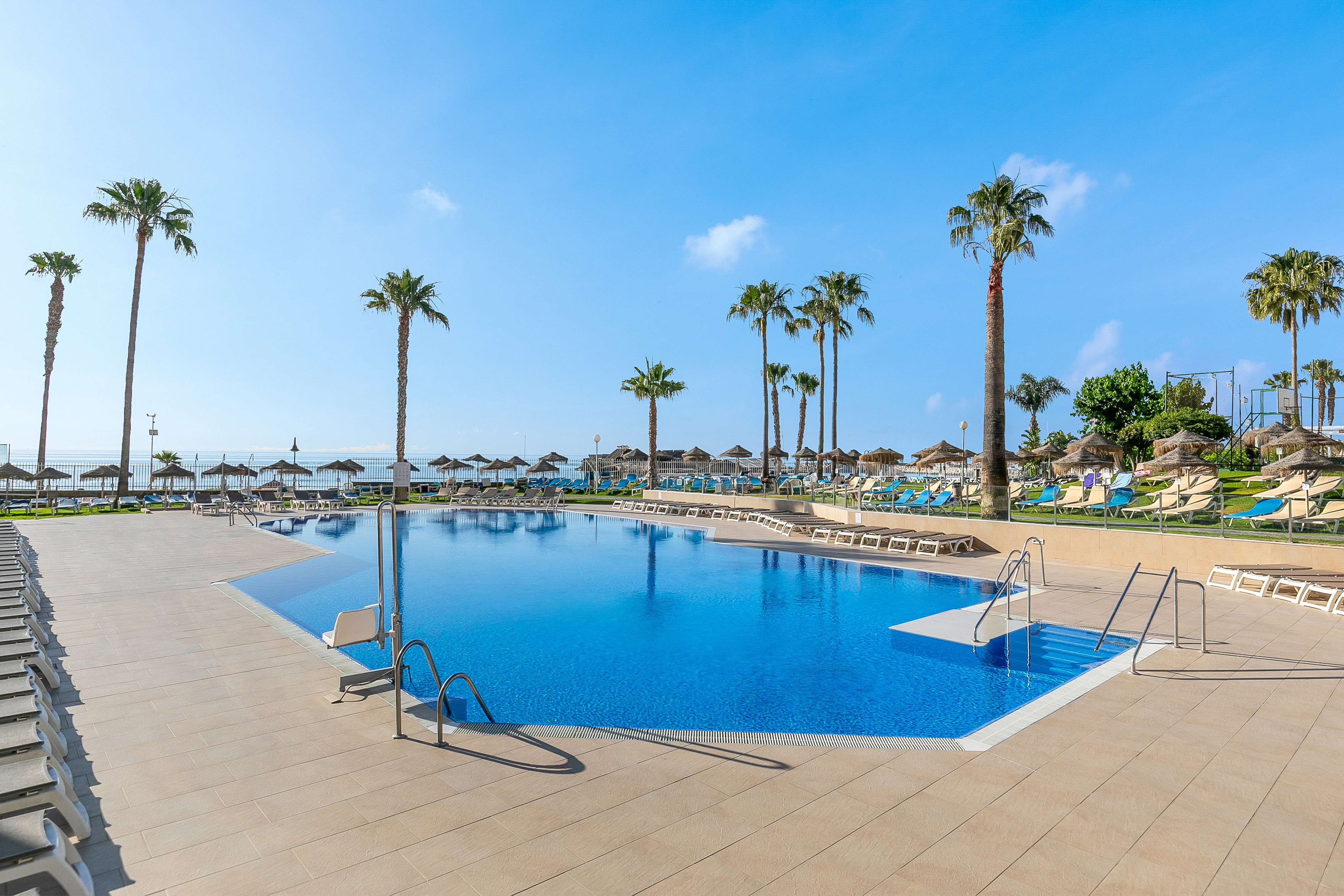 Tropical hotel pool, palm trees, sunbeds, and sea view under blue sky.
