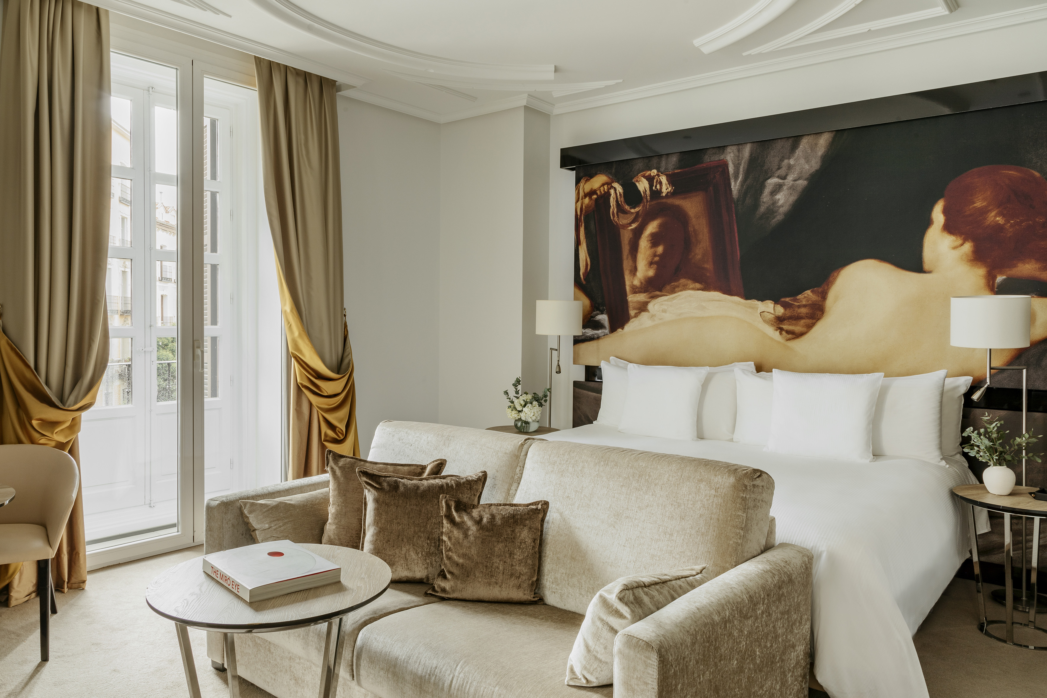 Elegant hotel room: large bed, sofa, classical art mural, balcony windows.