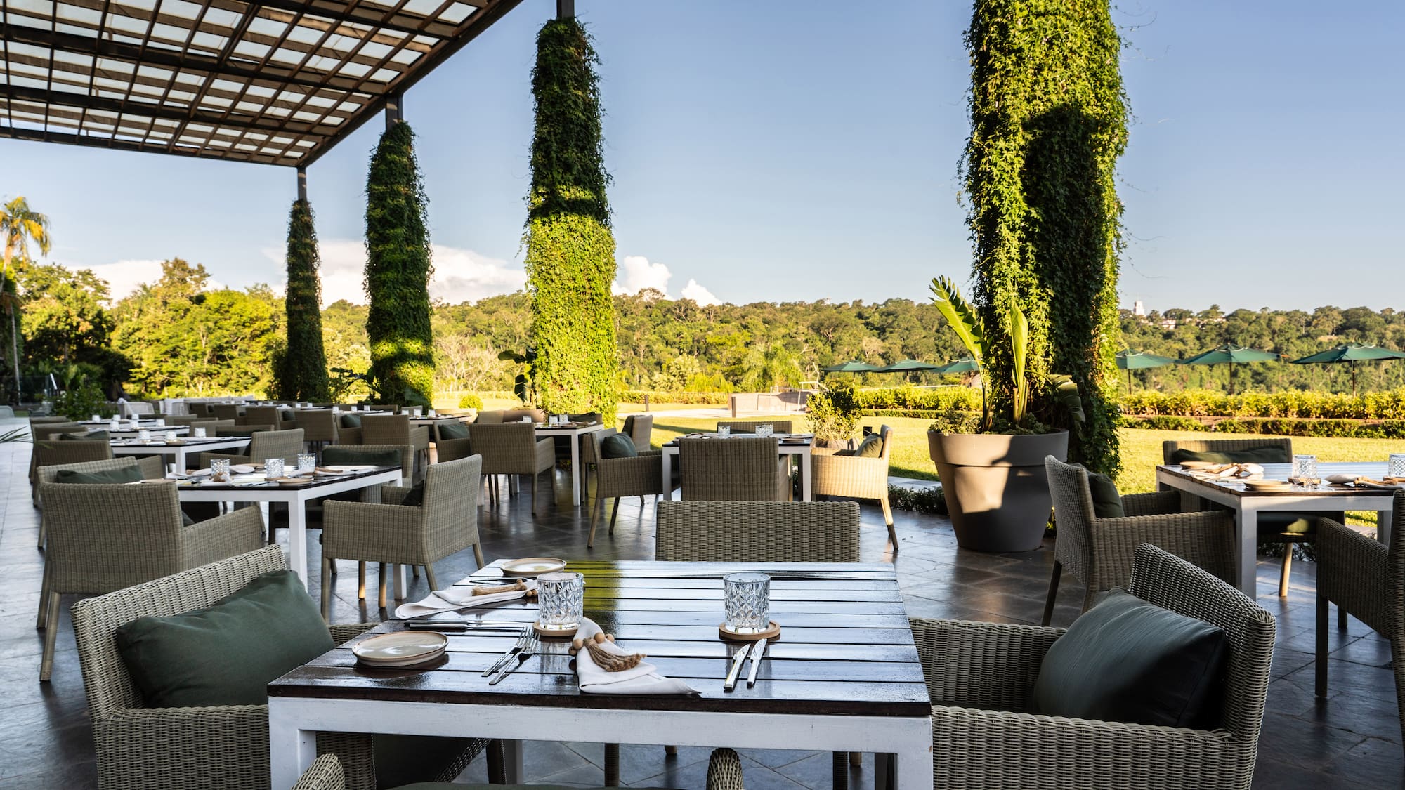 Restaurante terraza: mesas, sillas, columnas de hiedra, paisaje verde.