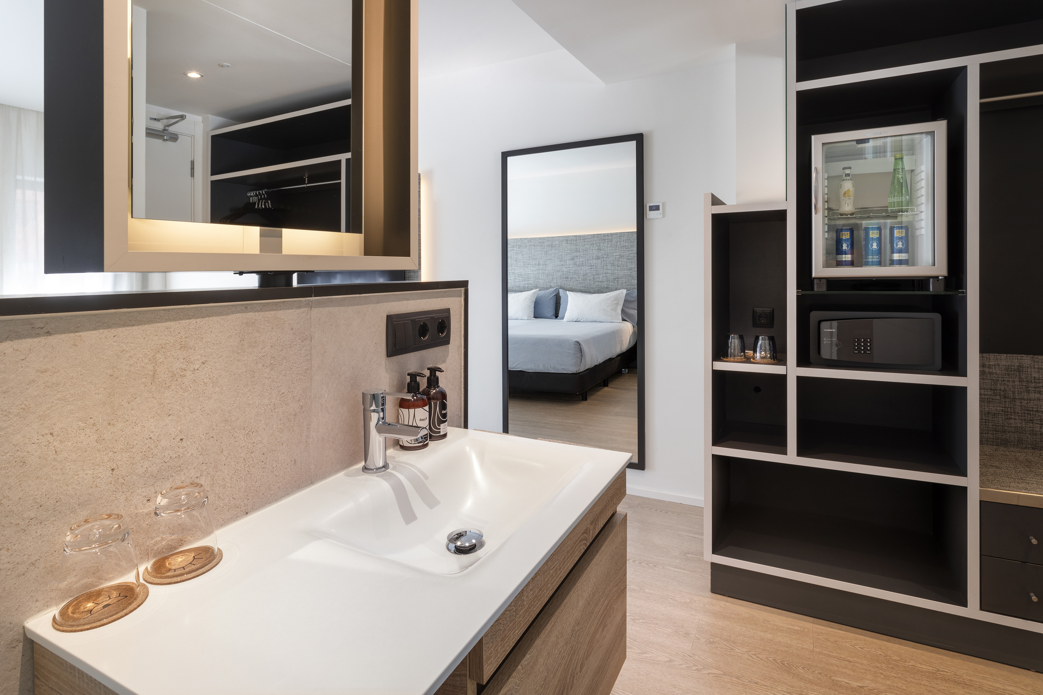Modern hotel room bathroom vanity with minibar and safe, reflecting bed.