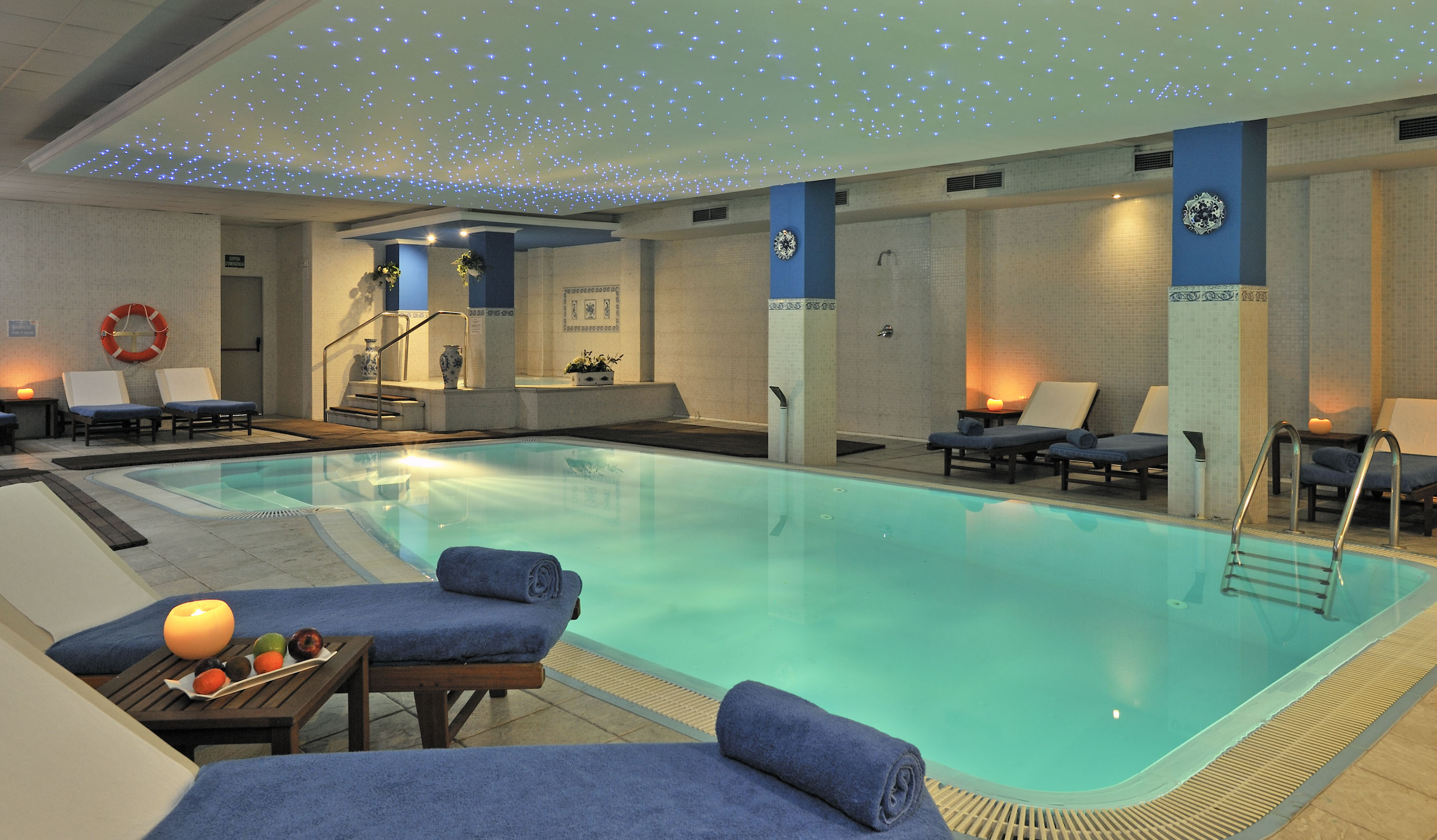 Indoor spa pool with blue lounge chairs, starry ceiling, and soft lighting.