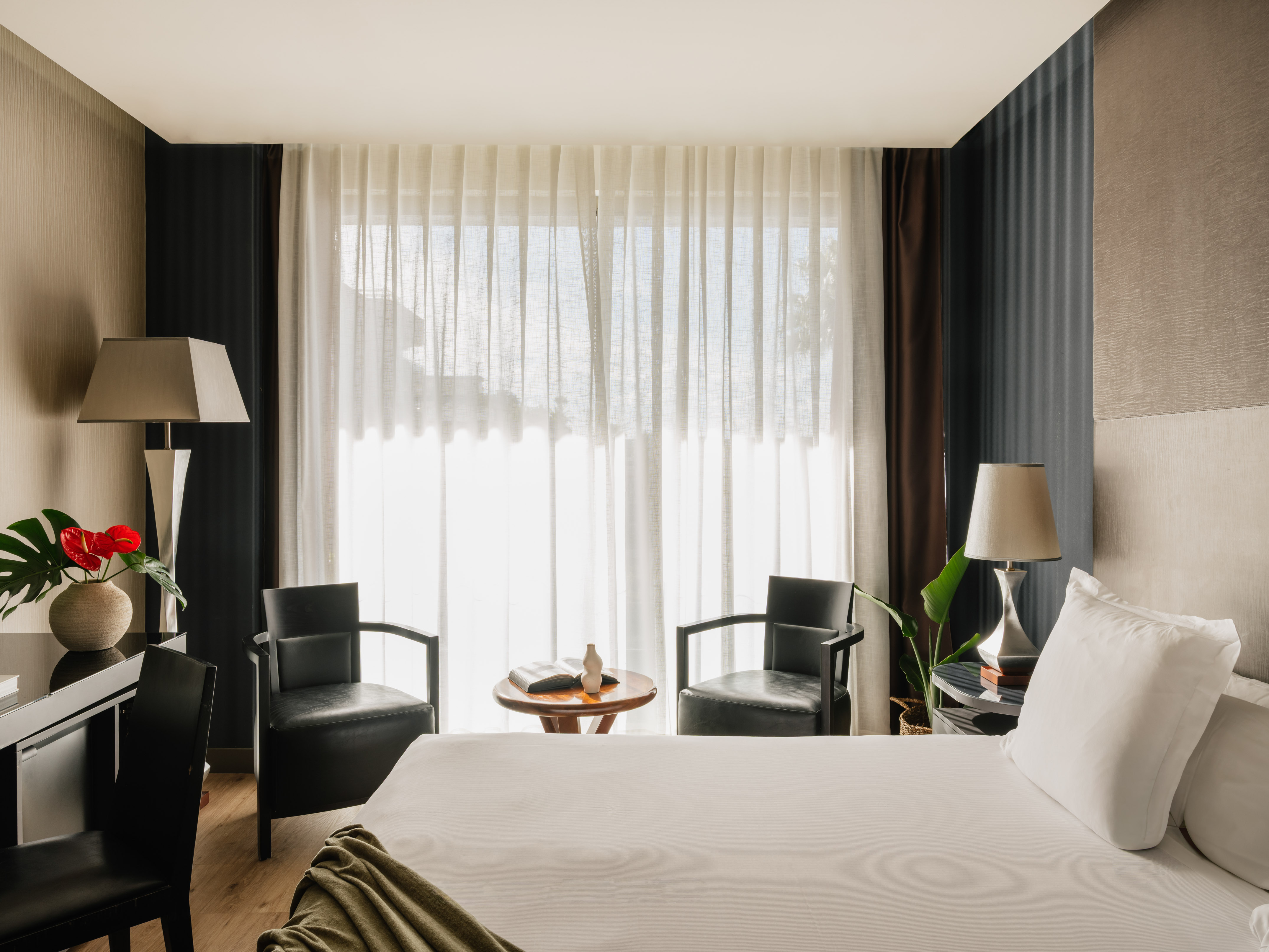 Modern hotel room with bed, armchairs, desk, and bright window.