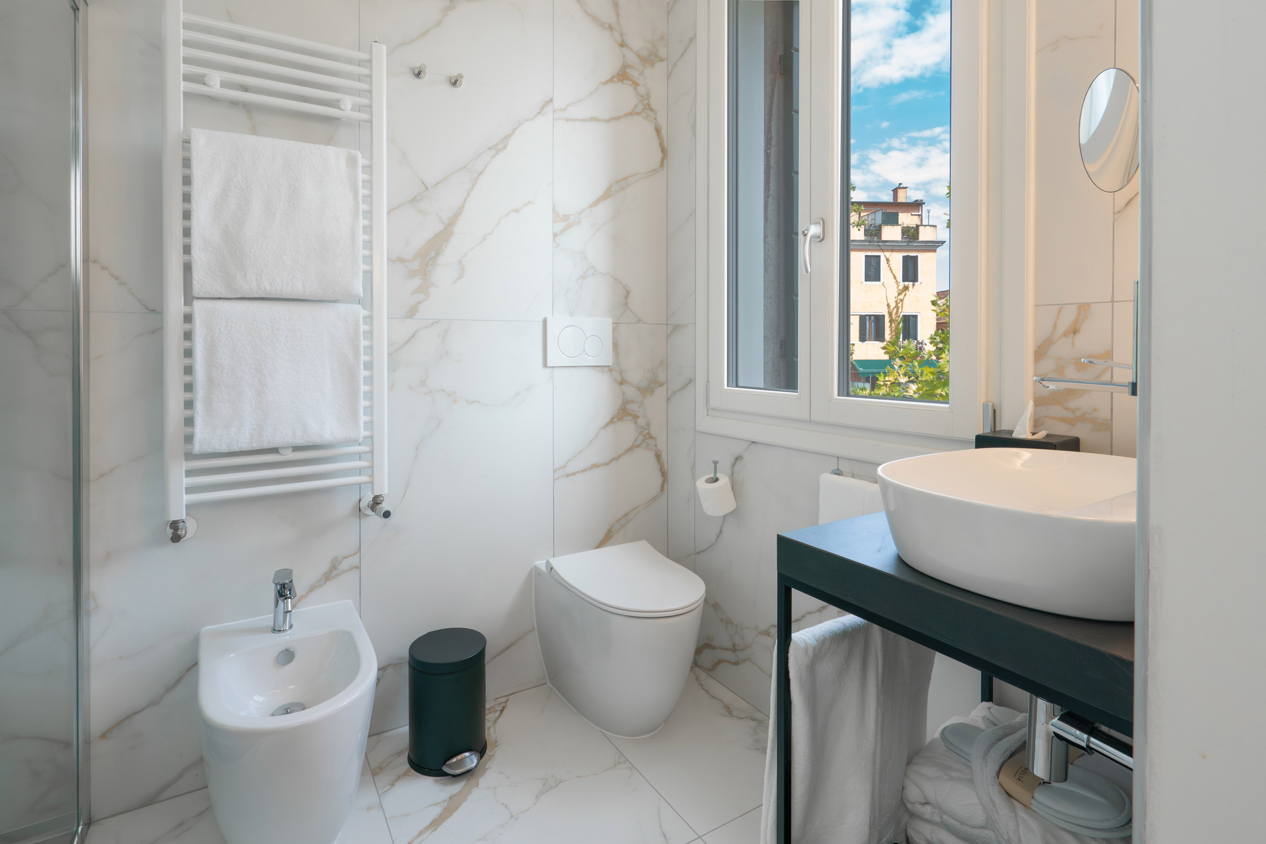 Modern white marble bathroom, toilet, bidet, sink, towel warmer, window.