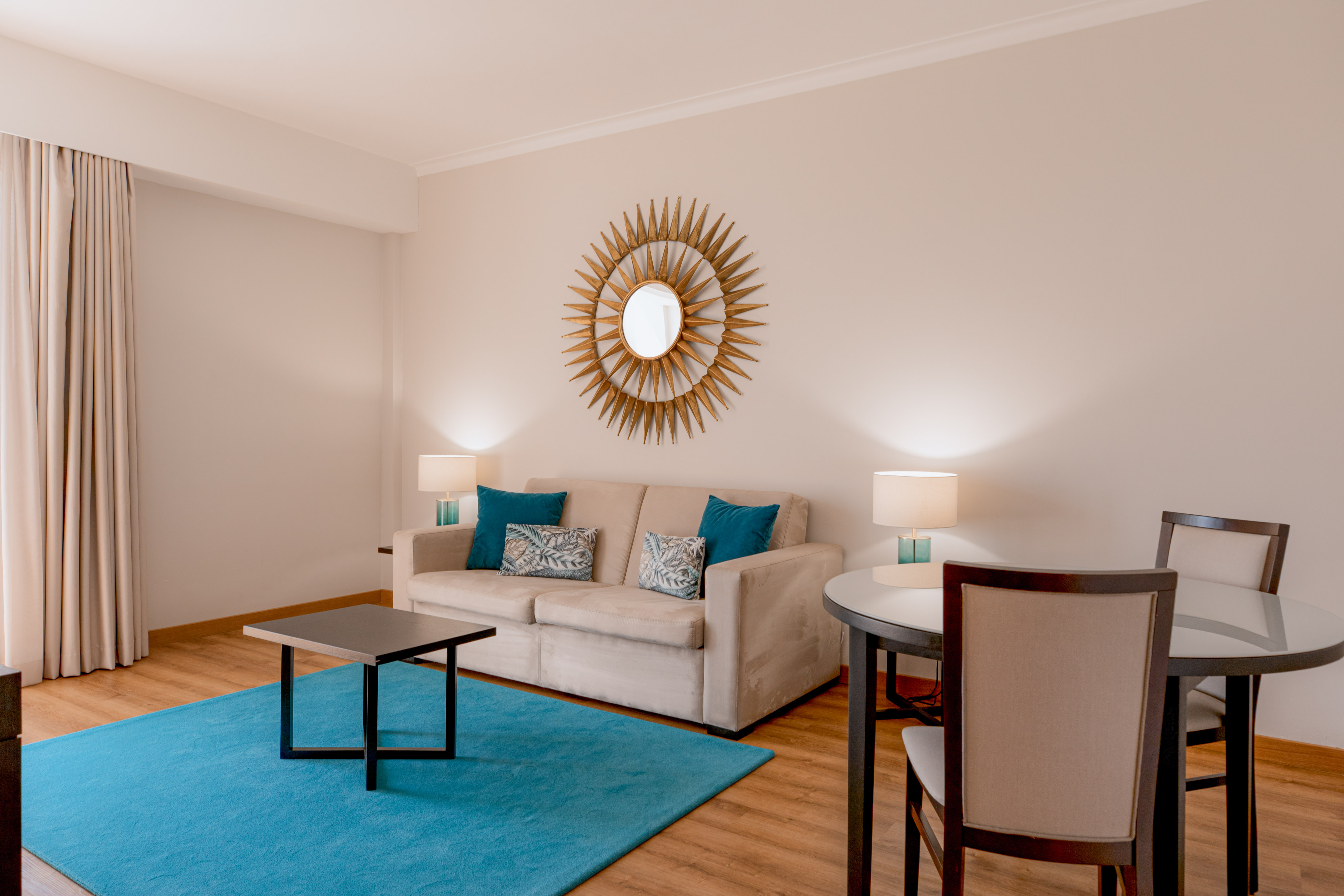 Modern living room with beige sofa, blue rug, sunburst mirror, and dining area.
