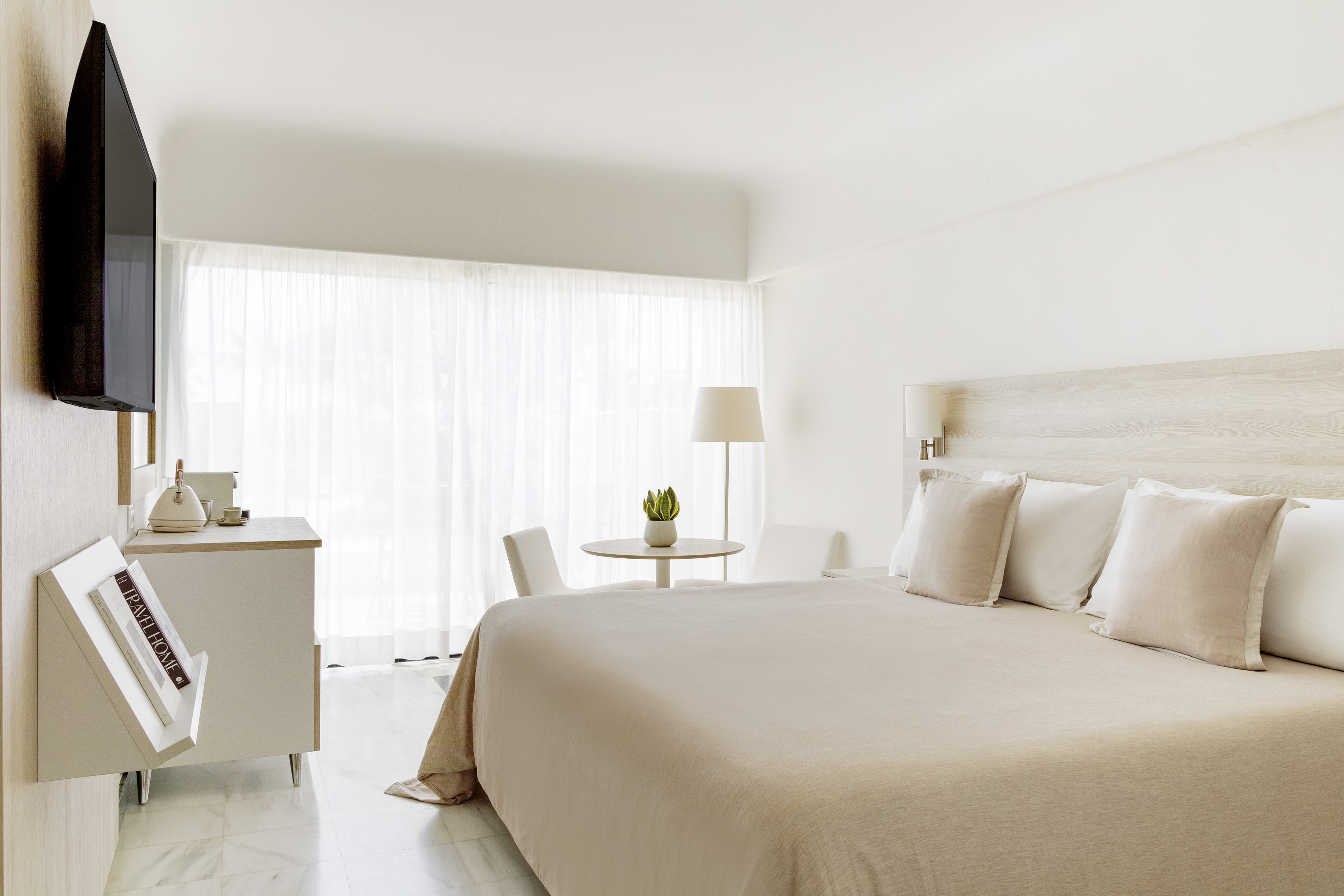 Modern hotel bedroom with king bed, neutral decor, TV, and bright window.