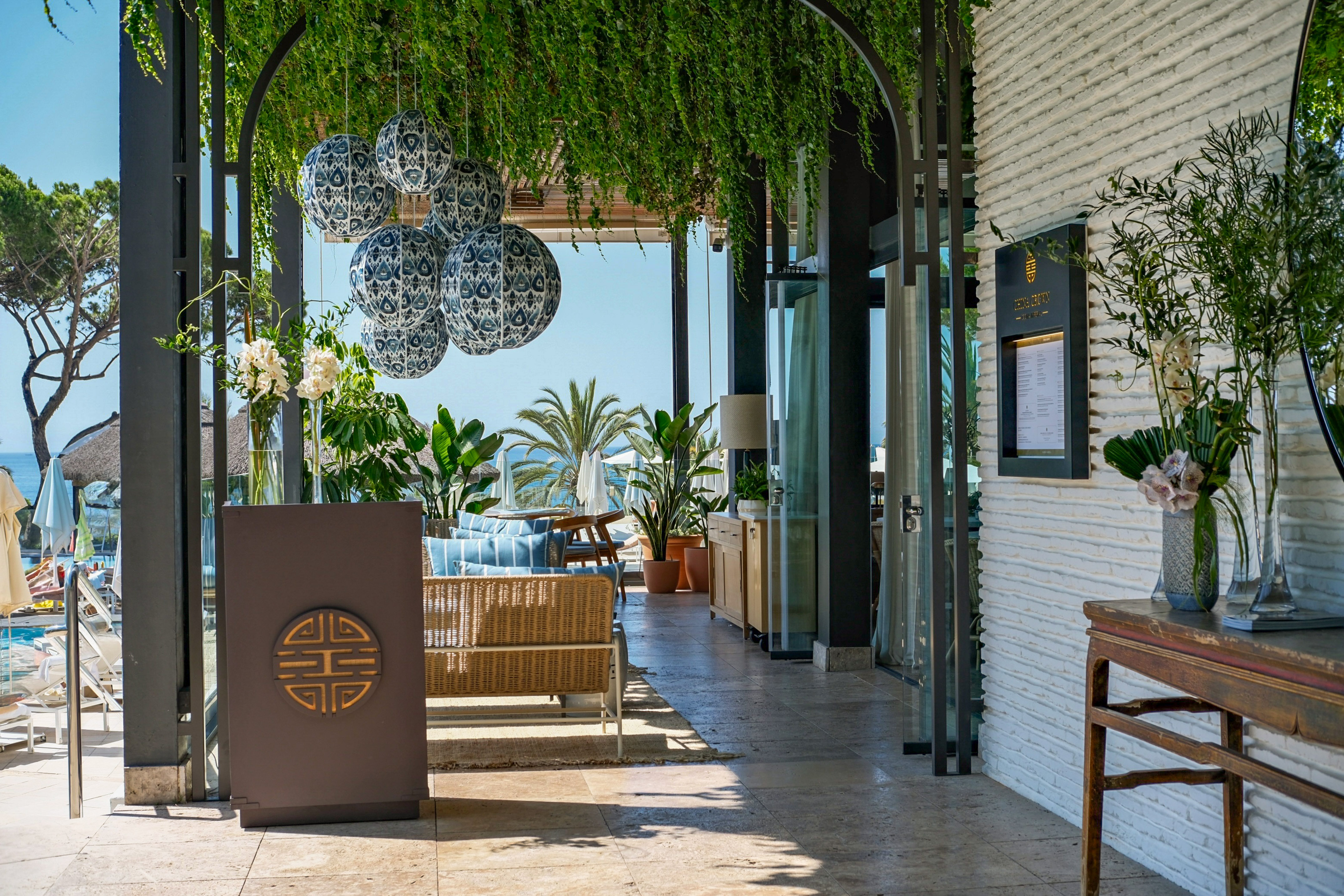 Outdoor restaurant patio with lush greenery, patterned lanterns, and ocean view.