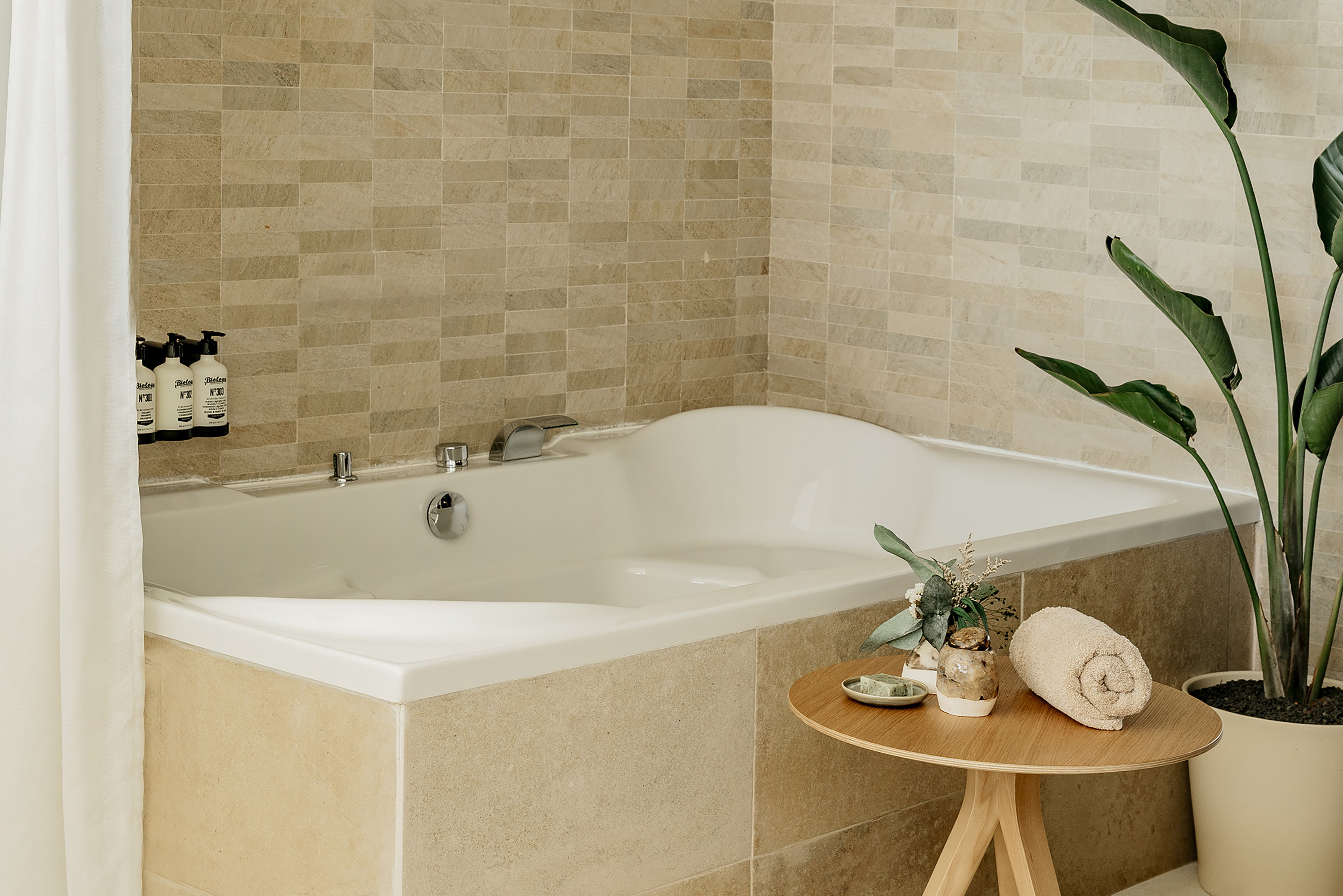 White corner bathtub in modern beige tiled bathroom with spa amenities.