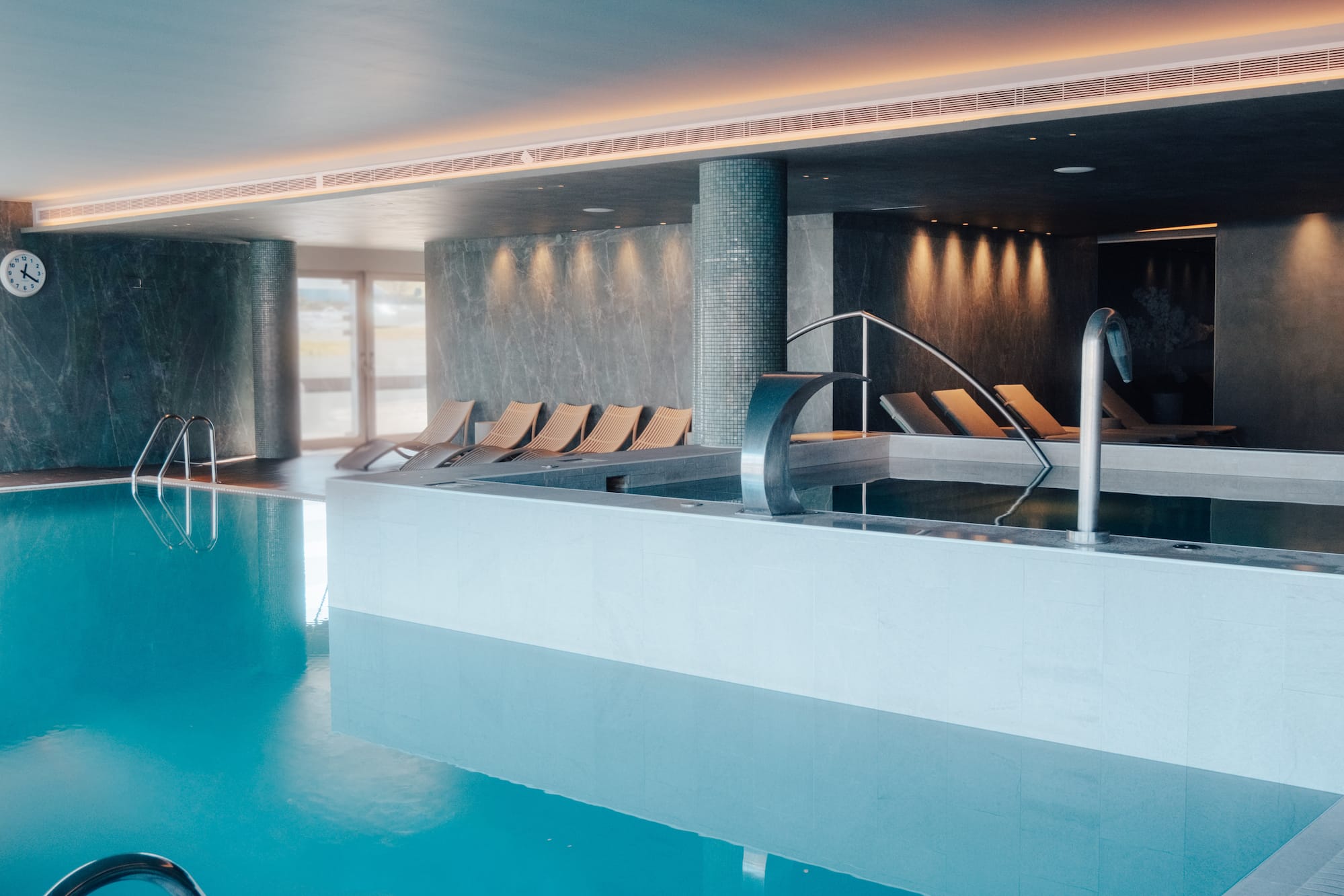 Modern indoor spa pool with blue water, lounge chairs, and a waterfall.