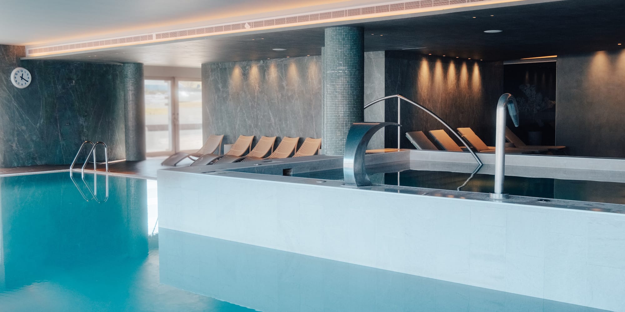 Luxury indoor pool area with blue water, lounge chairs, and a spa waterfall.