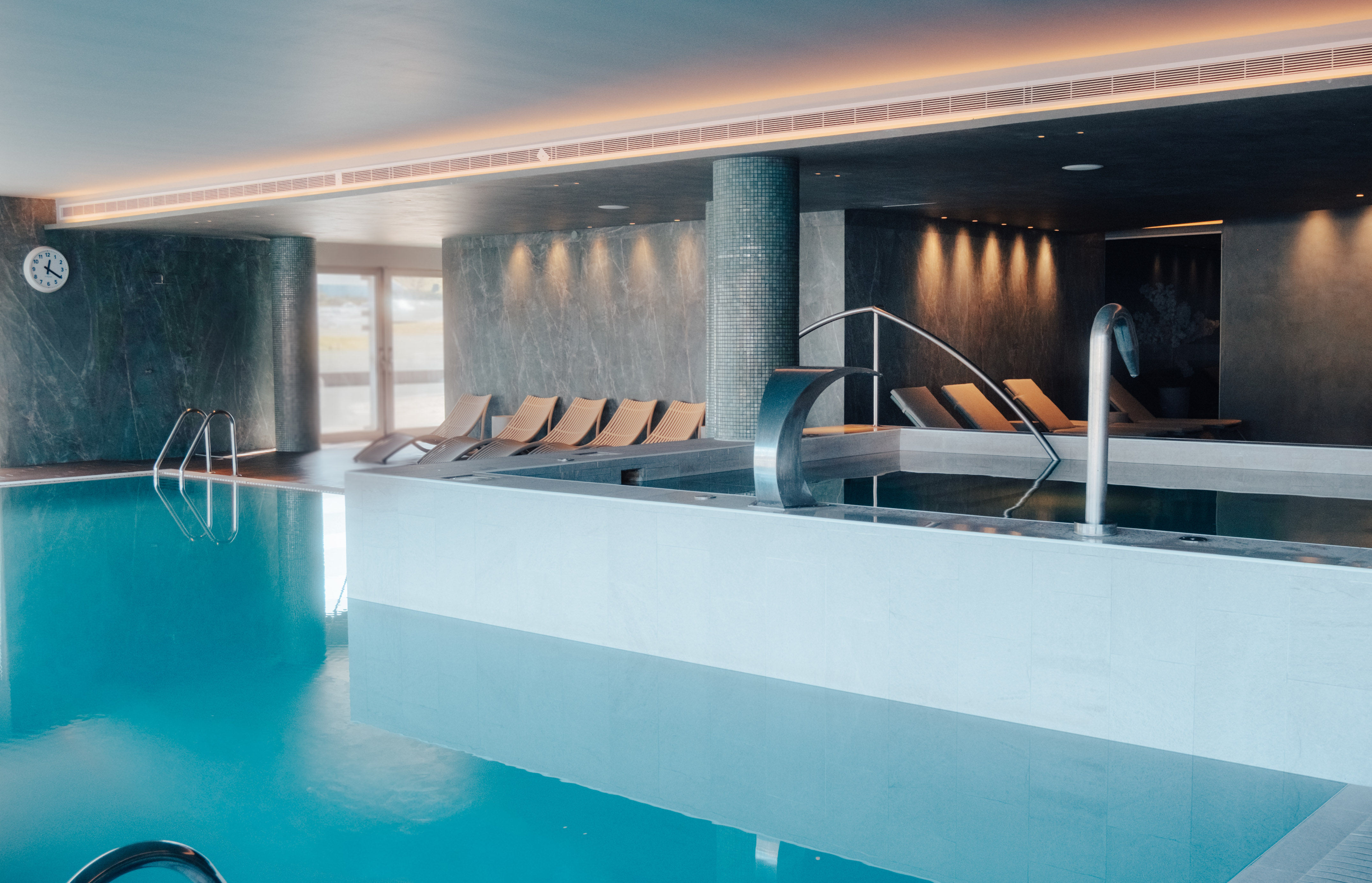 Luxury indoor pool area with blue water, lounge chairs, and a spa waterfall.