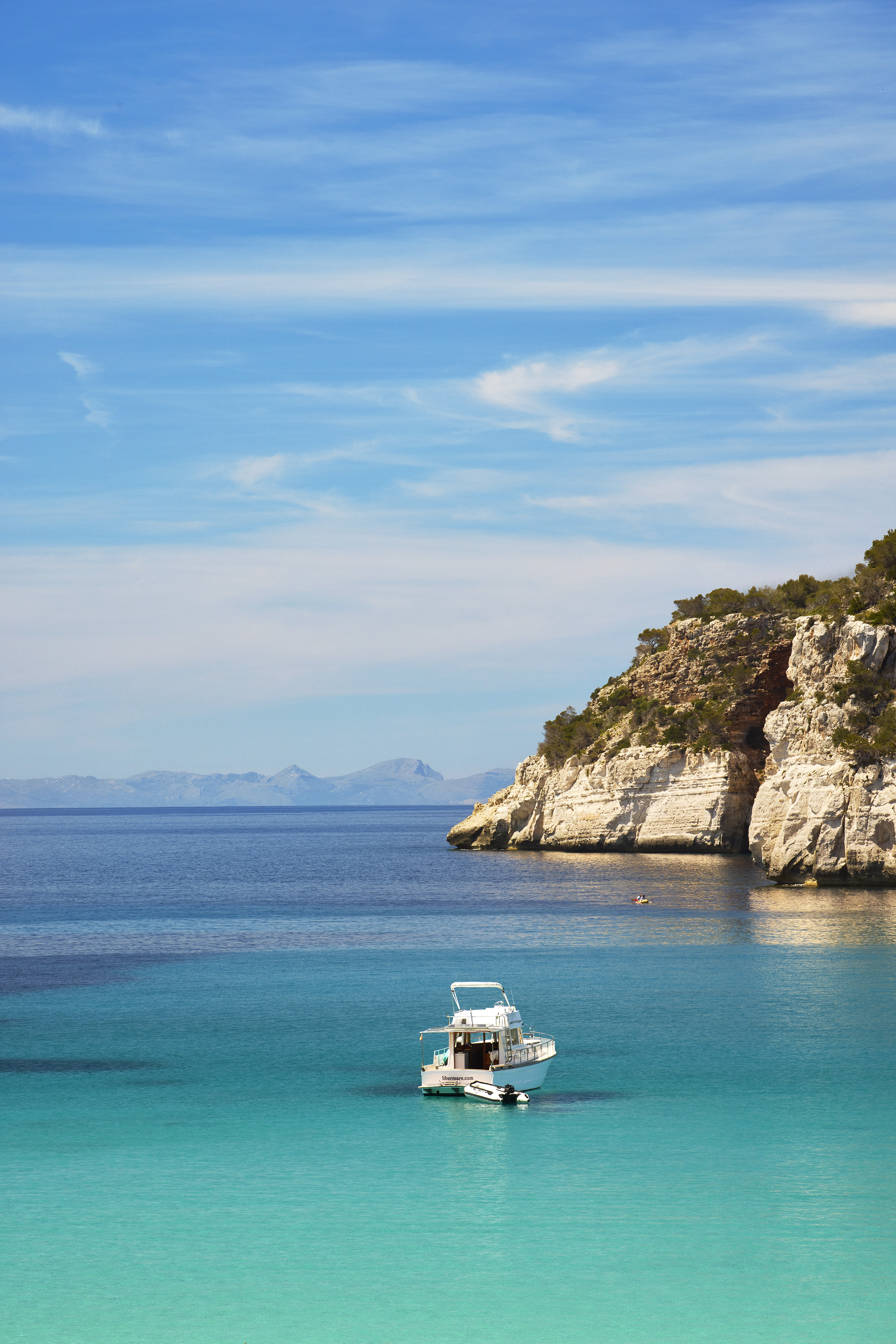 White yacht on vibrant turquoise water, rocky cliff, clear blue sky.