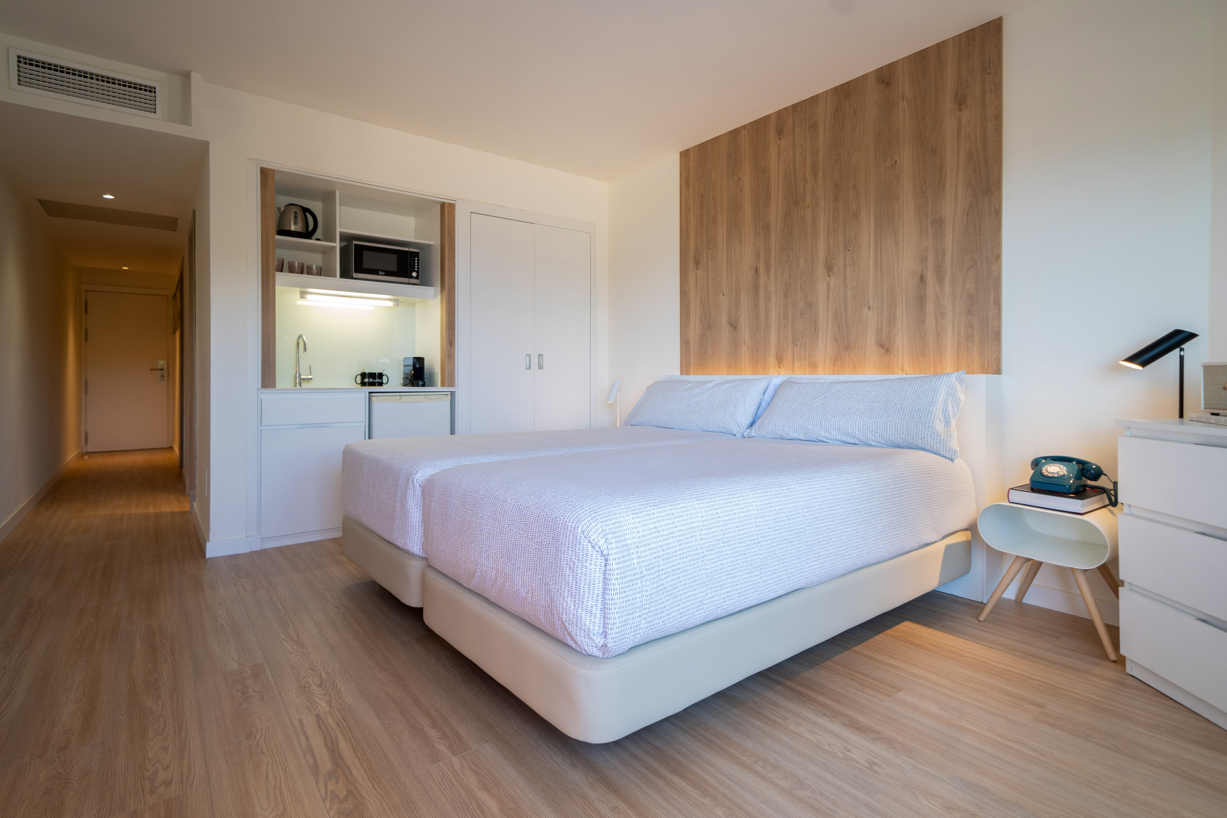 Minimalist hotel room with twin beds, kitchenette, and wood paneling.