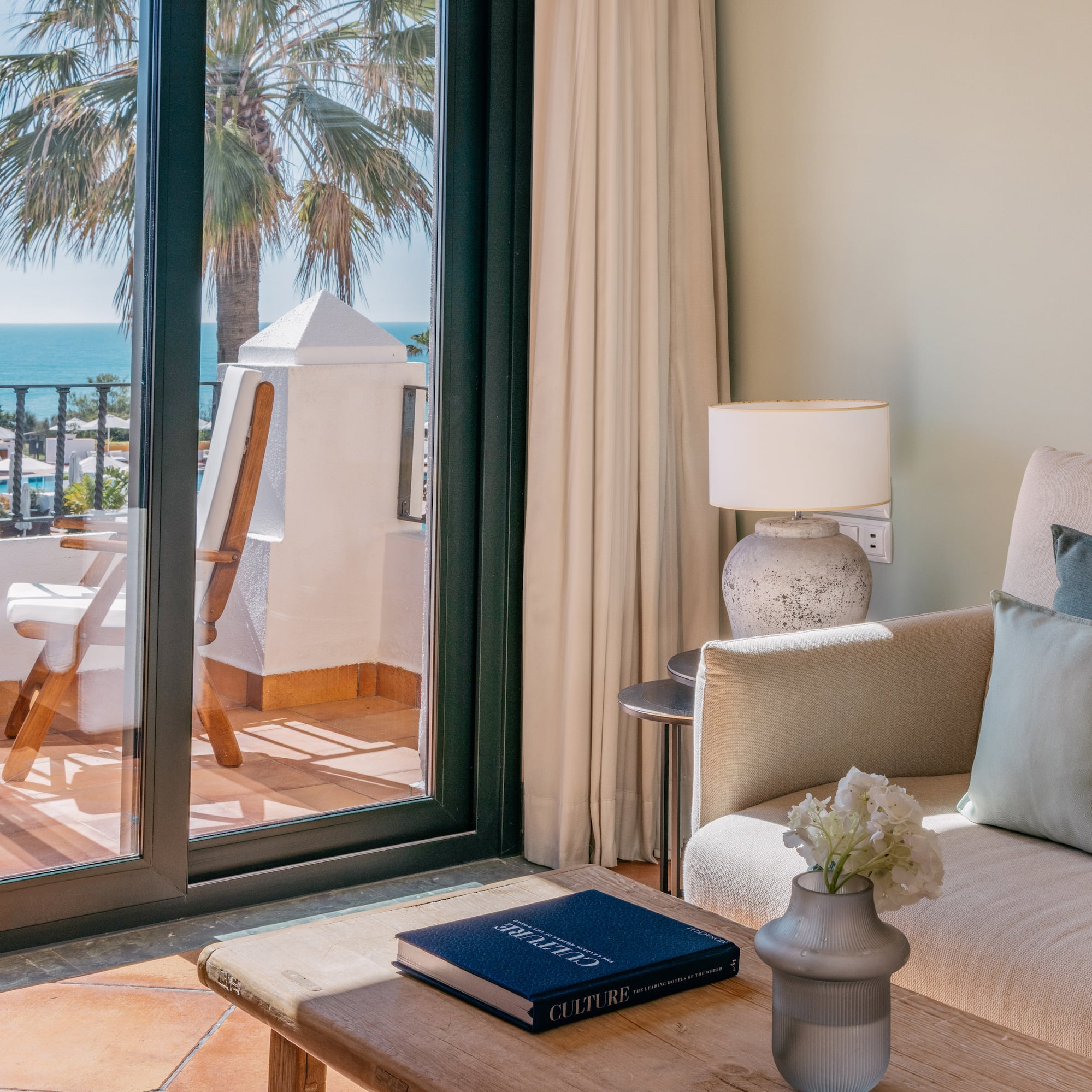 Luxury hotel suite with ocean view balcony, sofa, and palm tree.