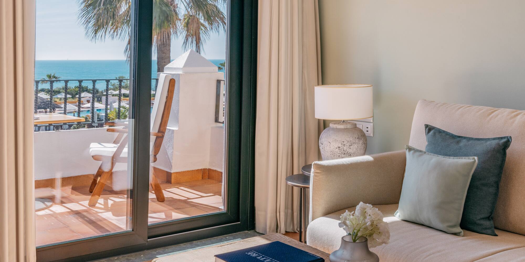 Luxury hotel suite with ocean view balcony, sofa, and palm tree.