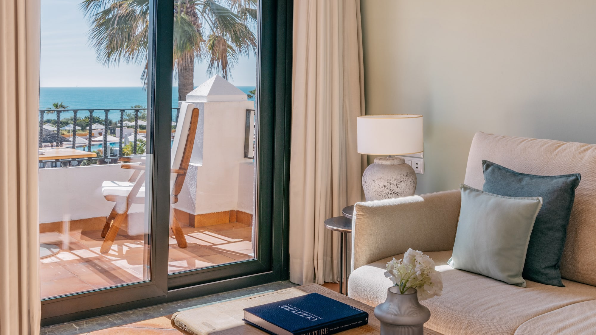 Luxury hotel suite with ocean view balcony, sofa, and palm tree.