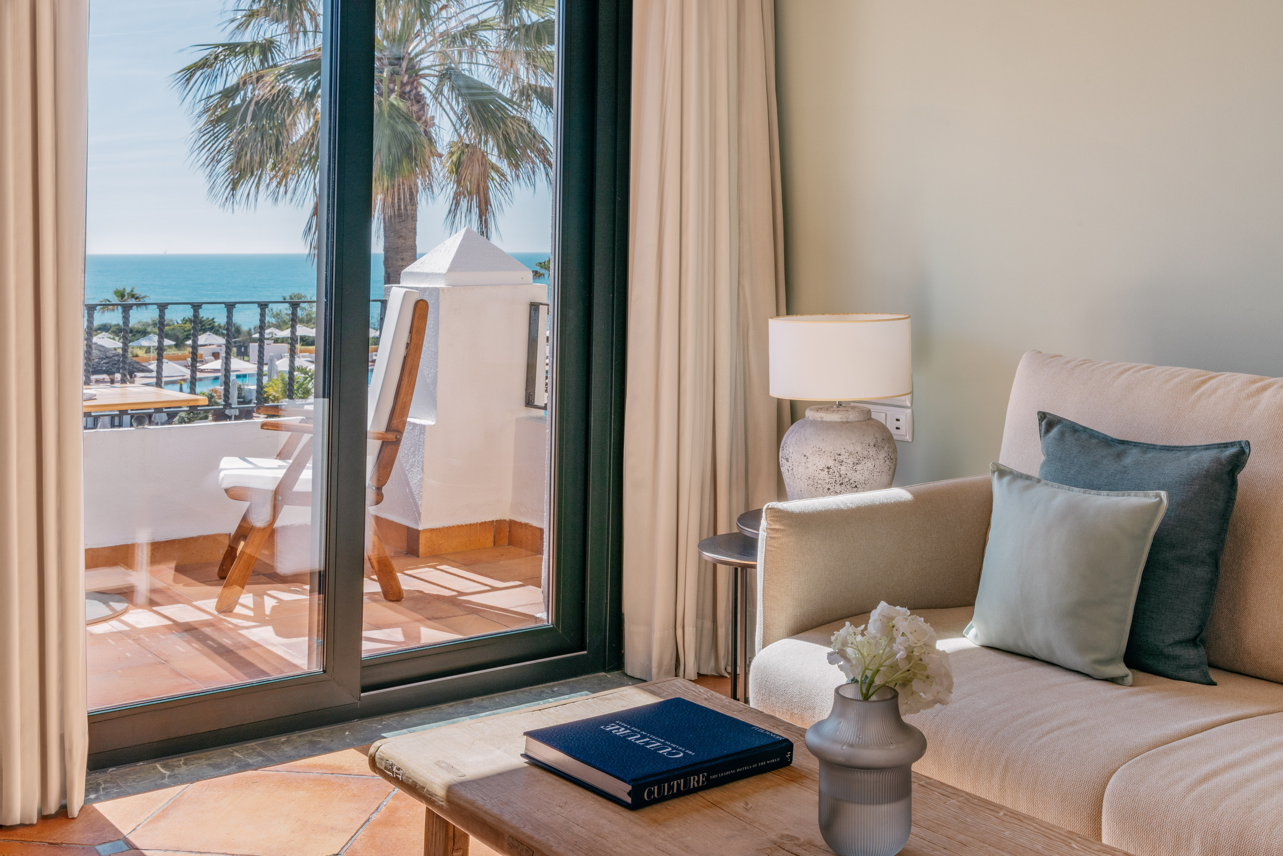 Luxury hotel suite with ocean view balcony, sofa, and palm tree.