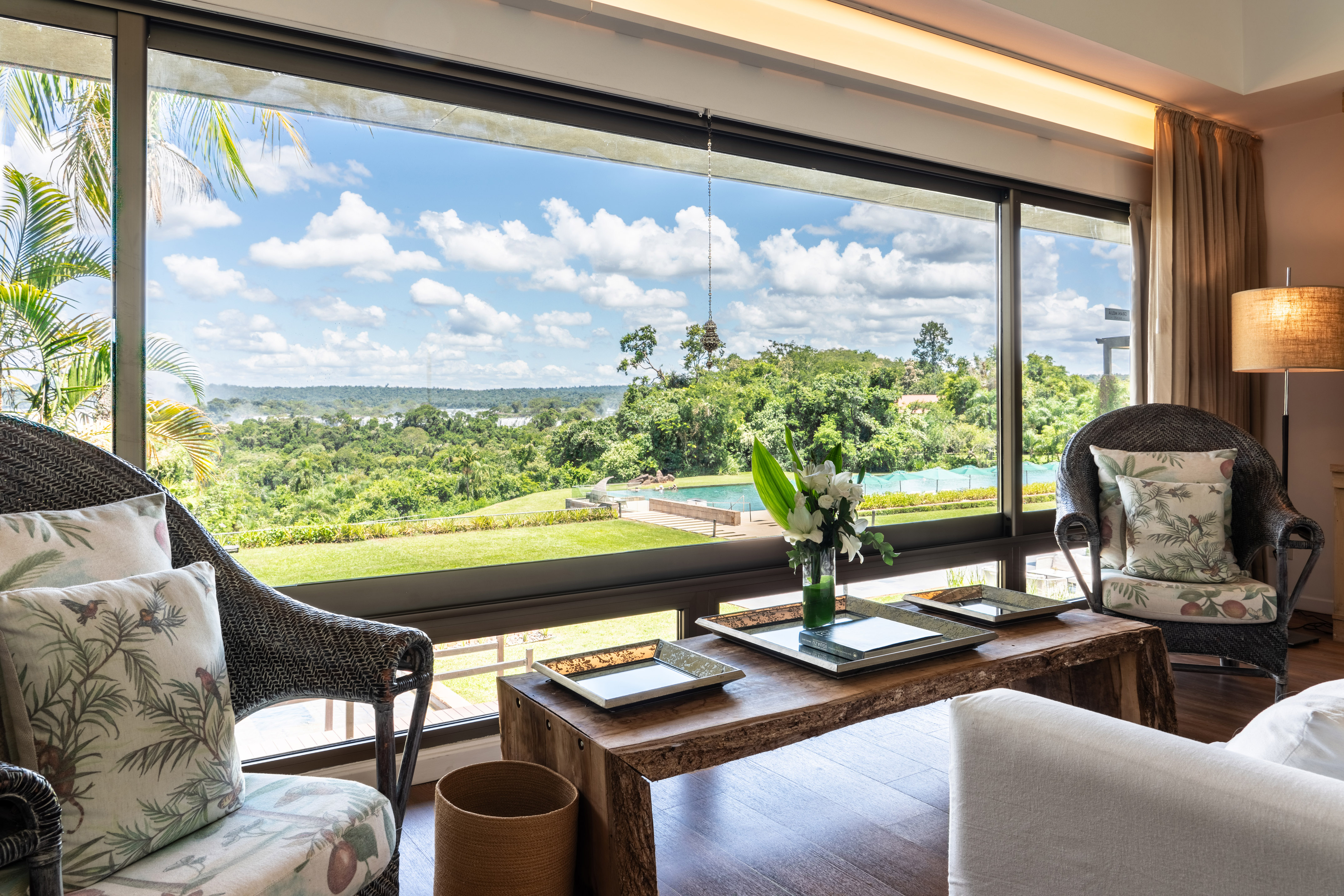 Tropical resort living room, panoramic jungle view, pool, and distant waterfalls.