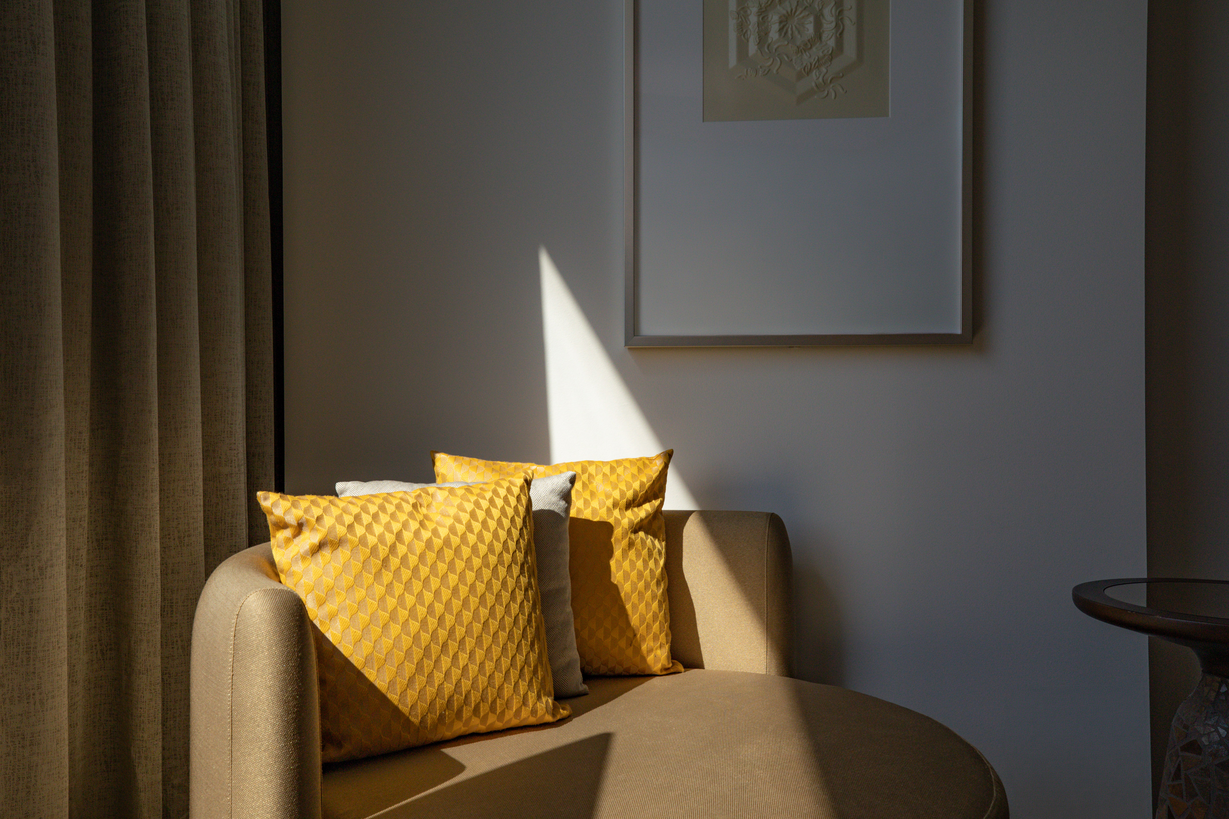 Sunlit chaise lounge with yellow geometric pillows, framed art, and drapes.