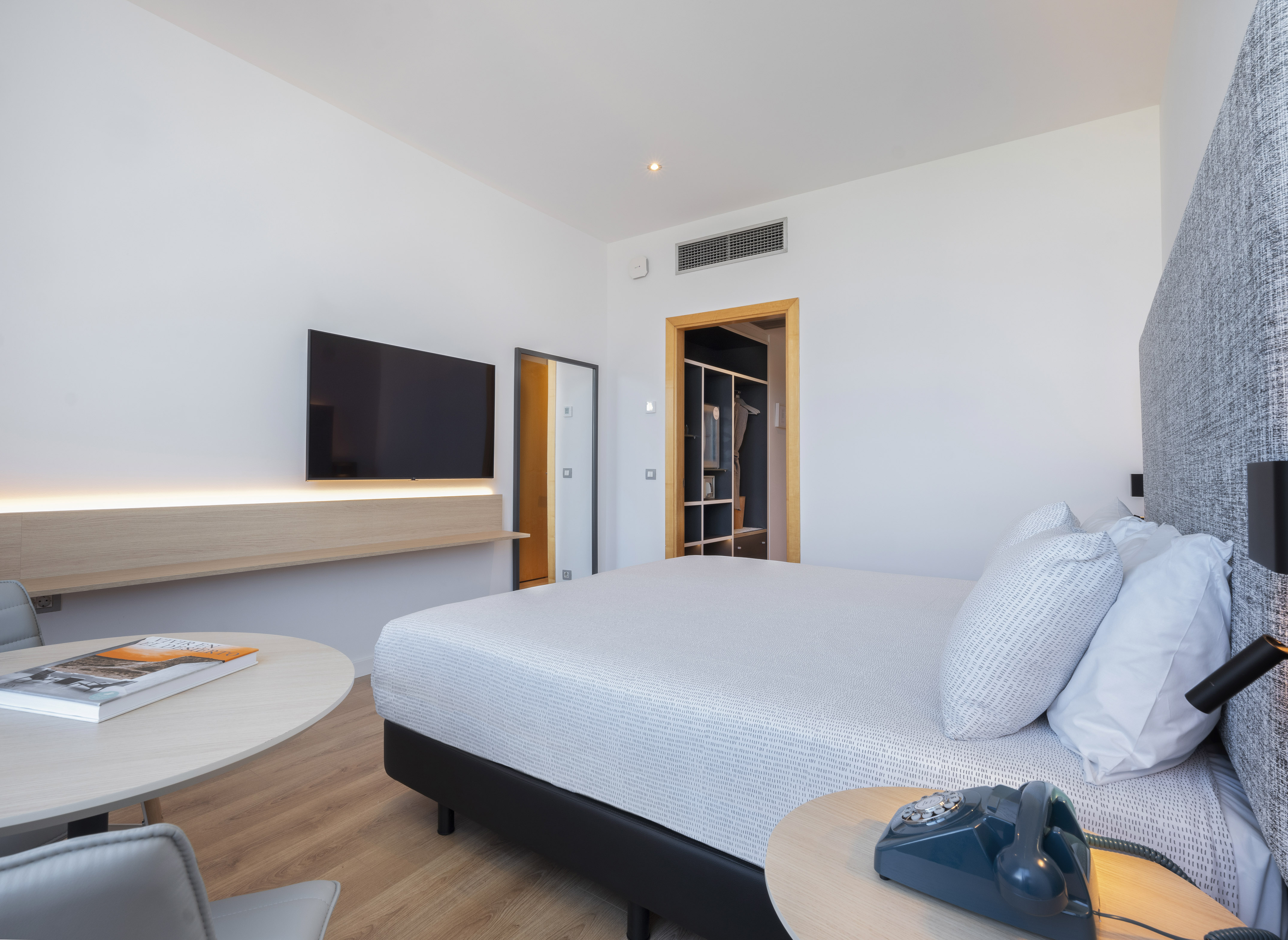 Modern hotel room with large bed, wall-mounted TV, light wood shelf, and open closet.