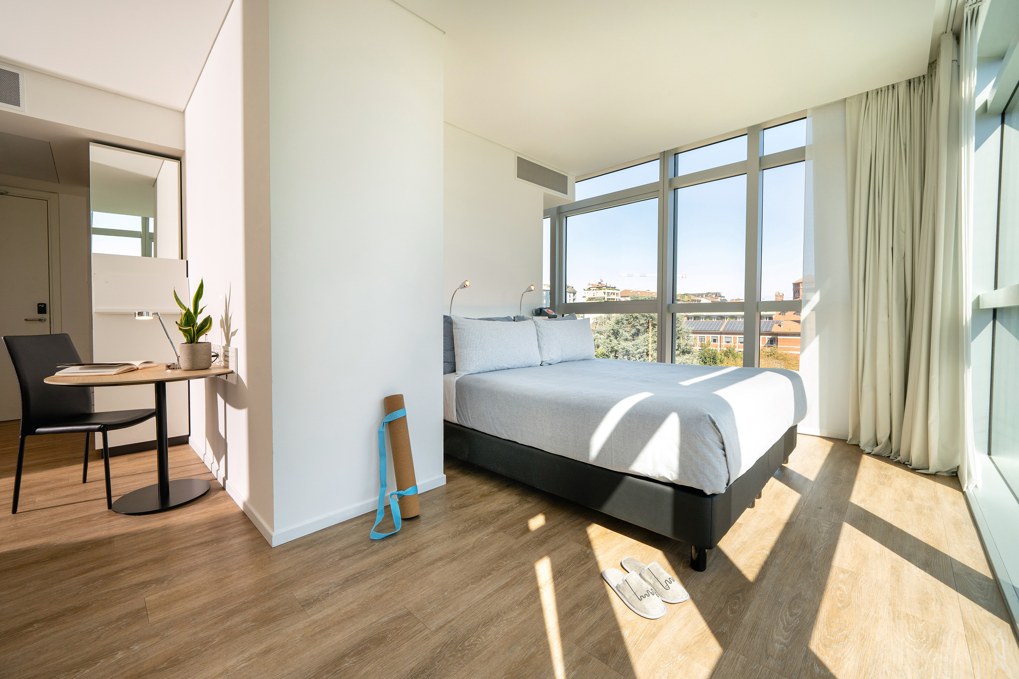 Modern bright bedroom with large windows, wooden floor, bed, desk, and yoga mat.