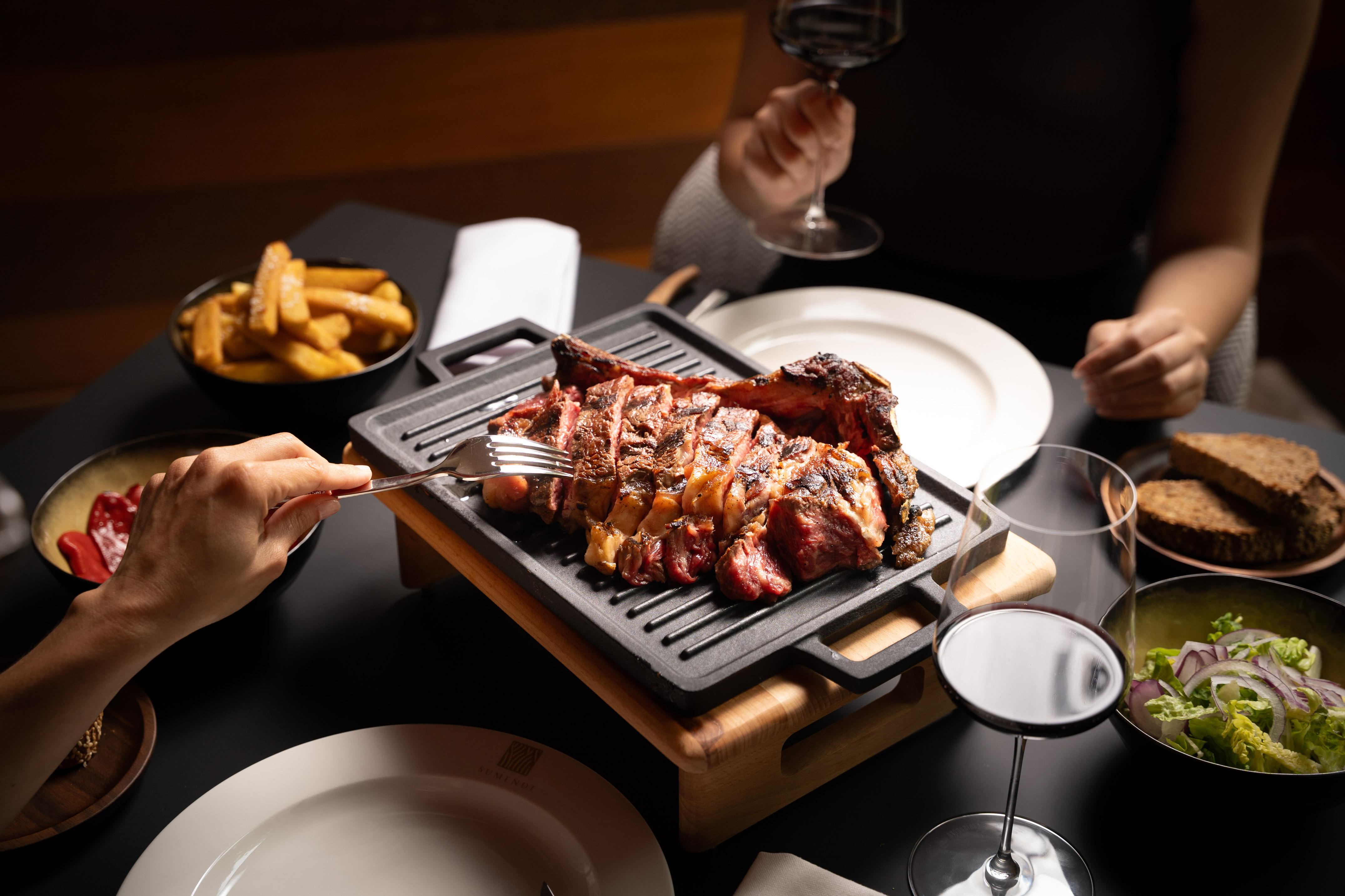 Diner forks juicy grilled steak, served with red wine, fries, and salad.
