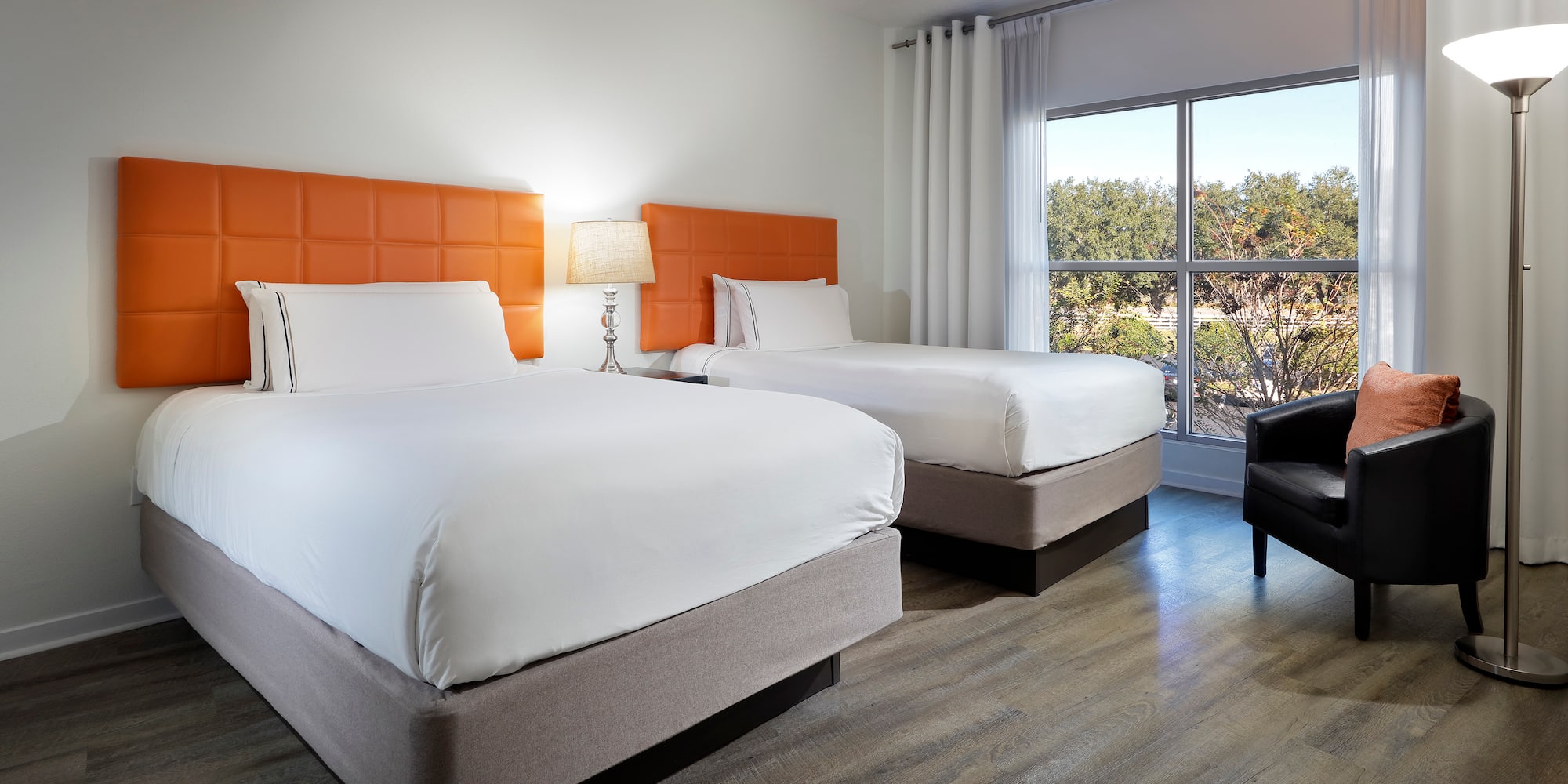 Hotel guest room with two double beds, orange headboards, window, armchair.
