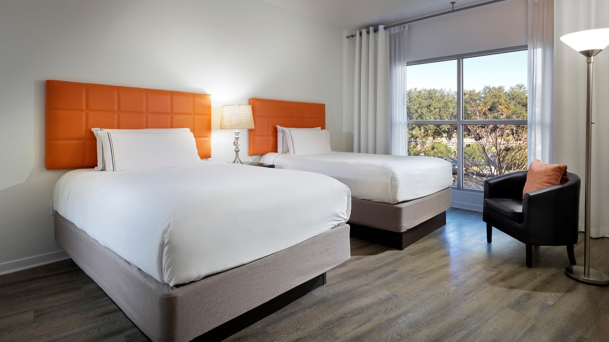 Hotel guest room with two double beds, orange headboards, window, armchair.