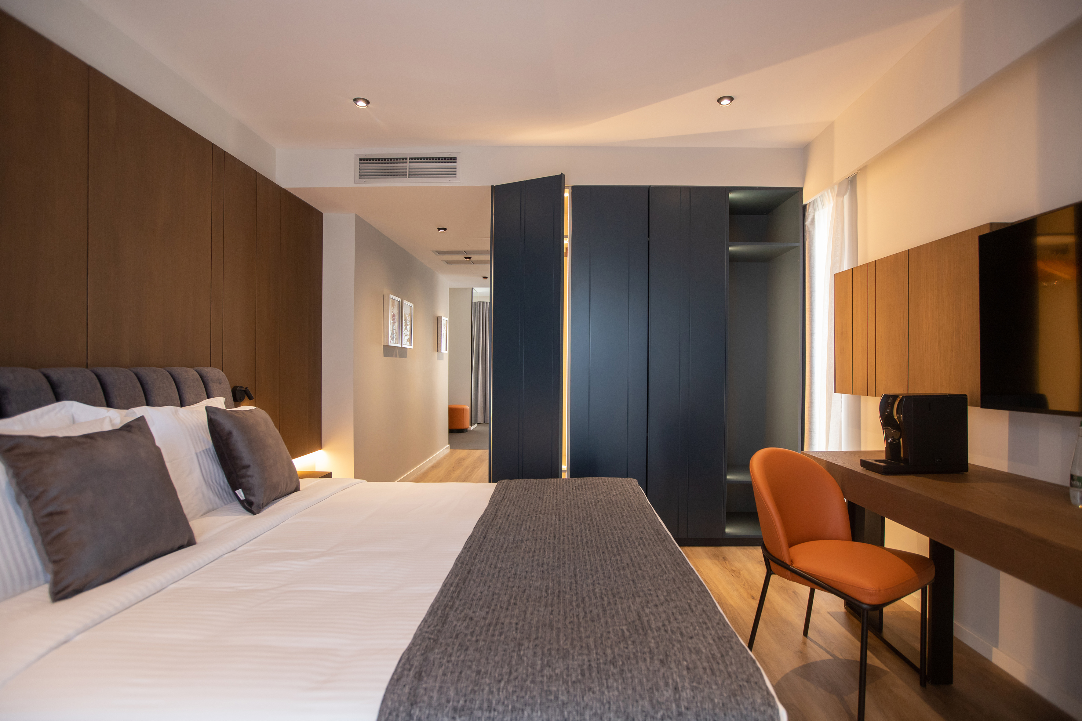 Modern hotel room: bed, dark wardrobe, desk with coffee machine, orange chair.