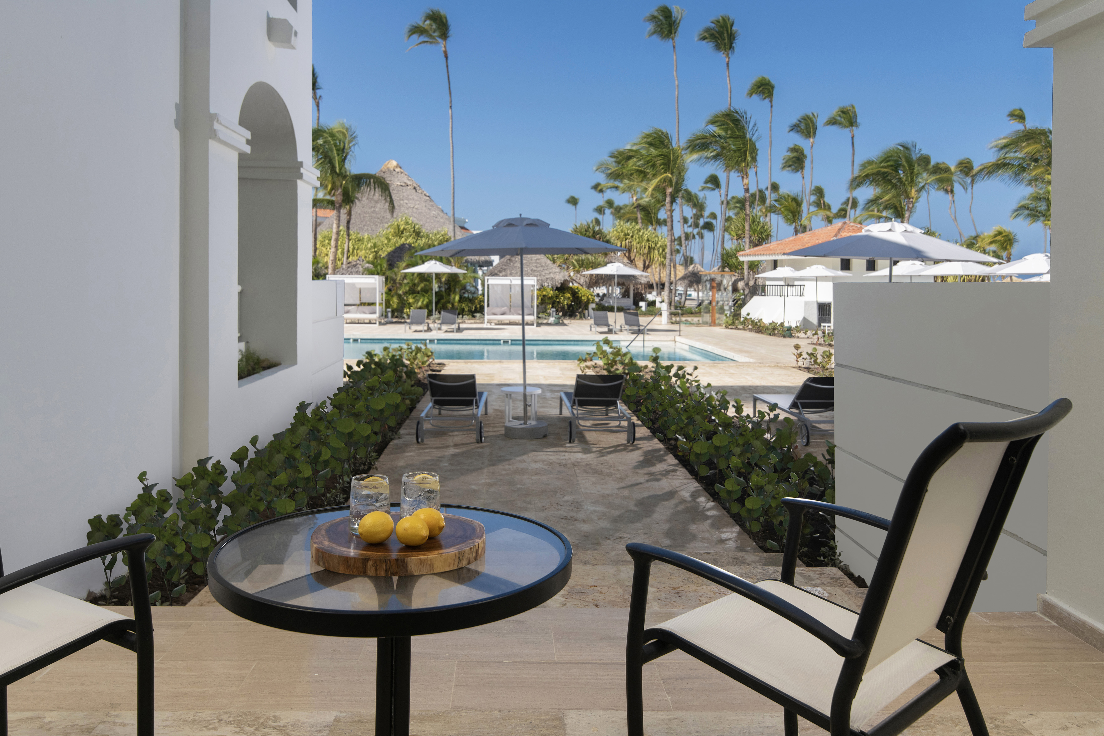 Tropical resort patio overlooking pool with drinks, palm trees, and sun loungers.