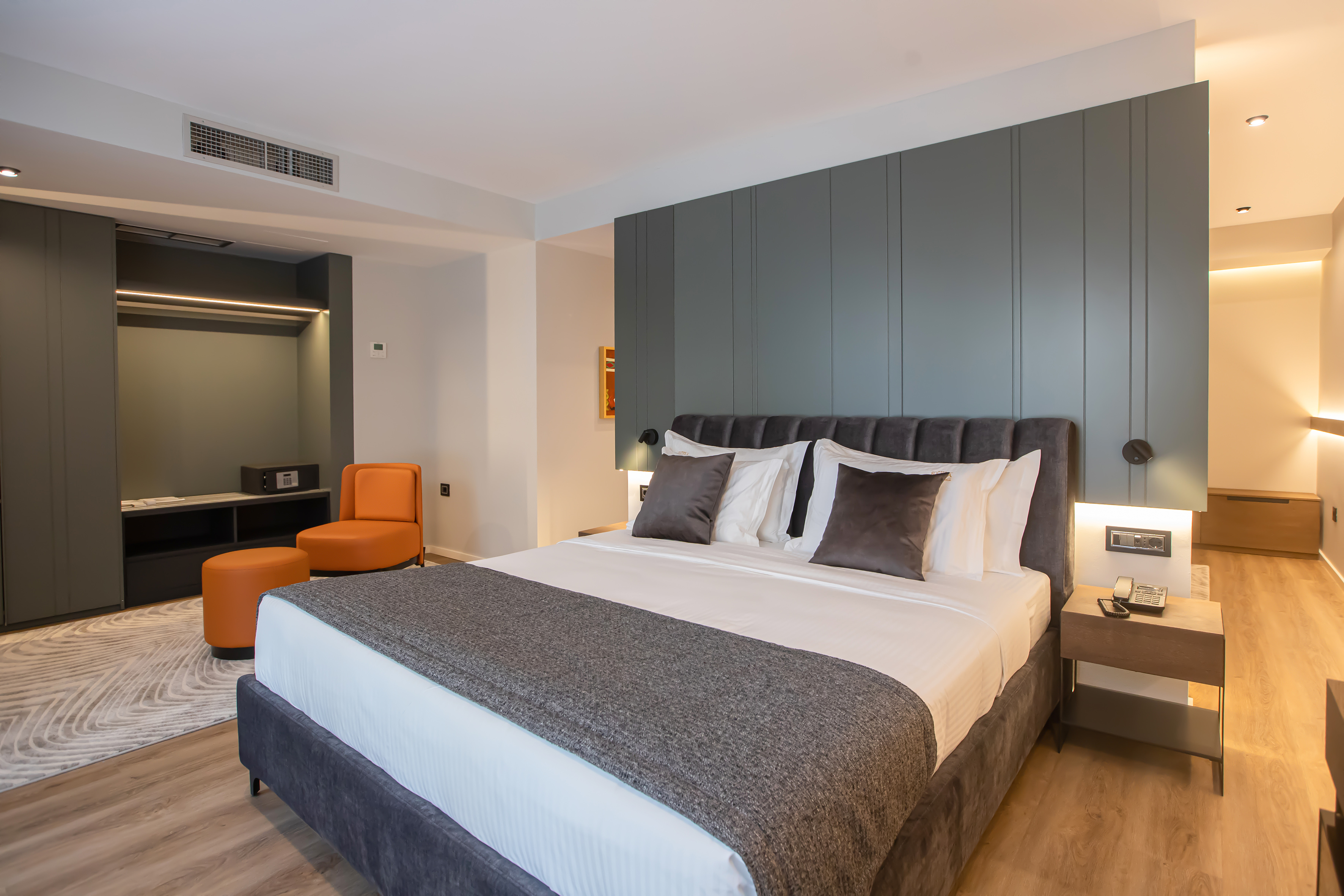 Modern hotel bedroom with king bed, grey decor, orange chair, and wood flooring.